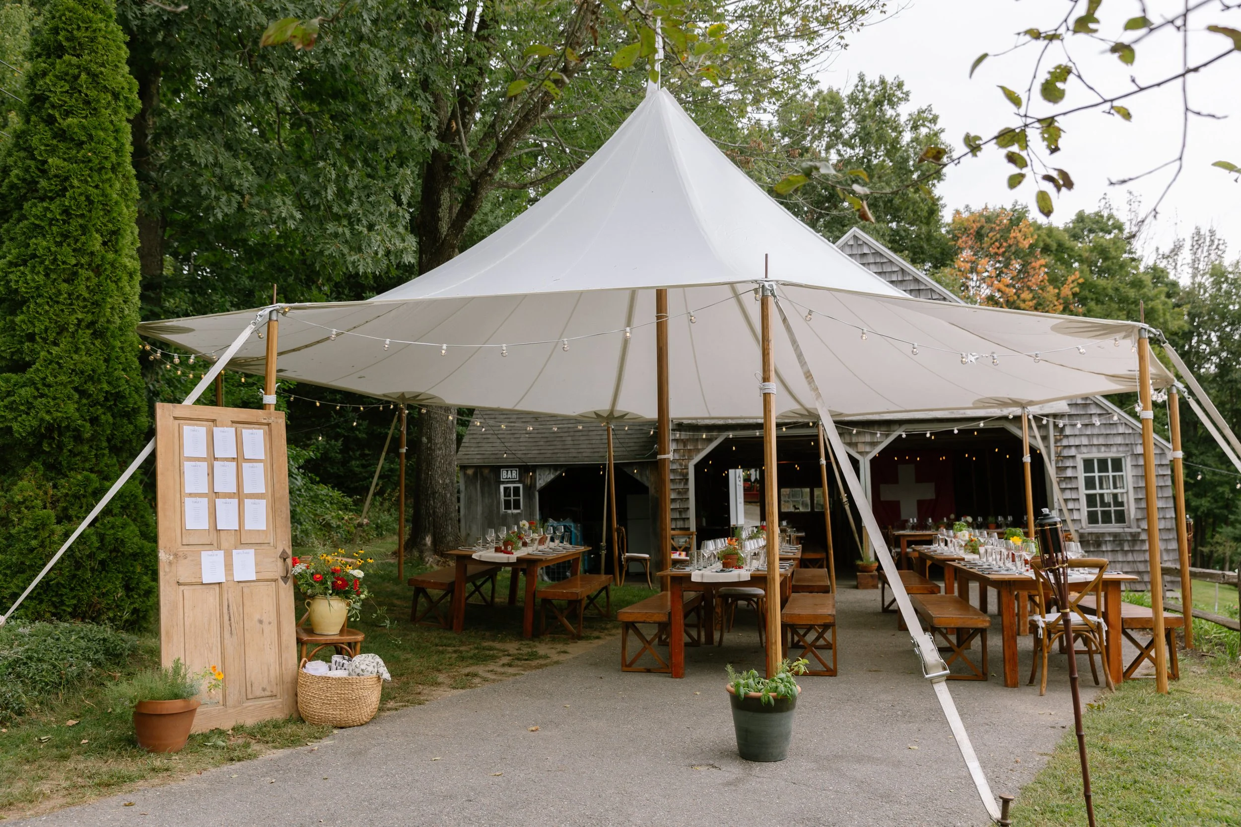 Outdoor wedding or event setup under a large white tent with wooden tables and chairs, decorated with tableware and glassware, surrounded by potted plants and greenery.