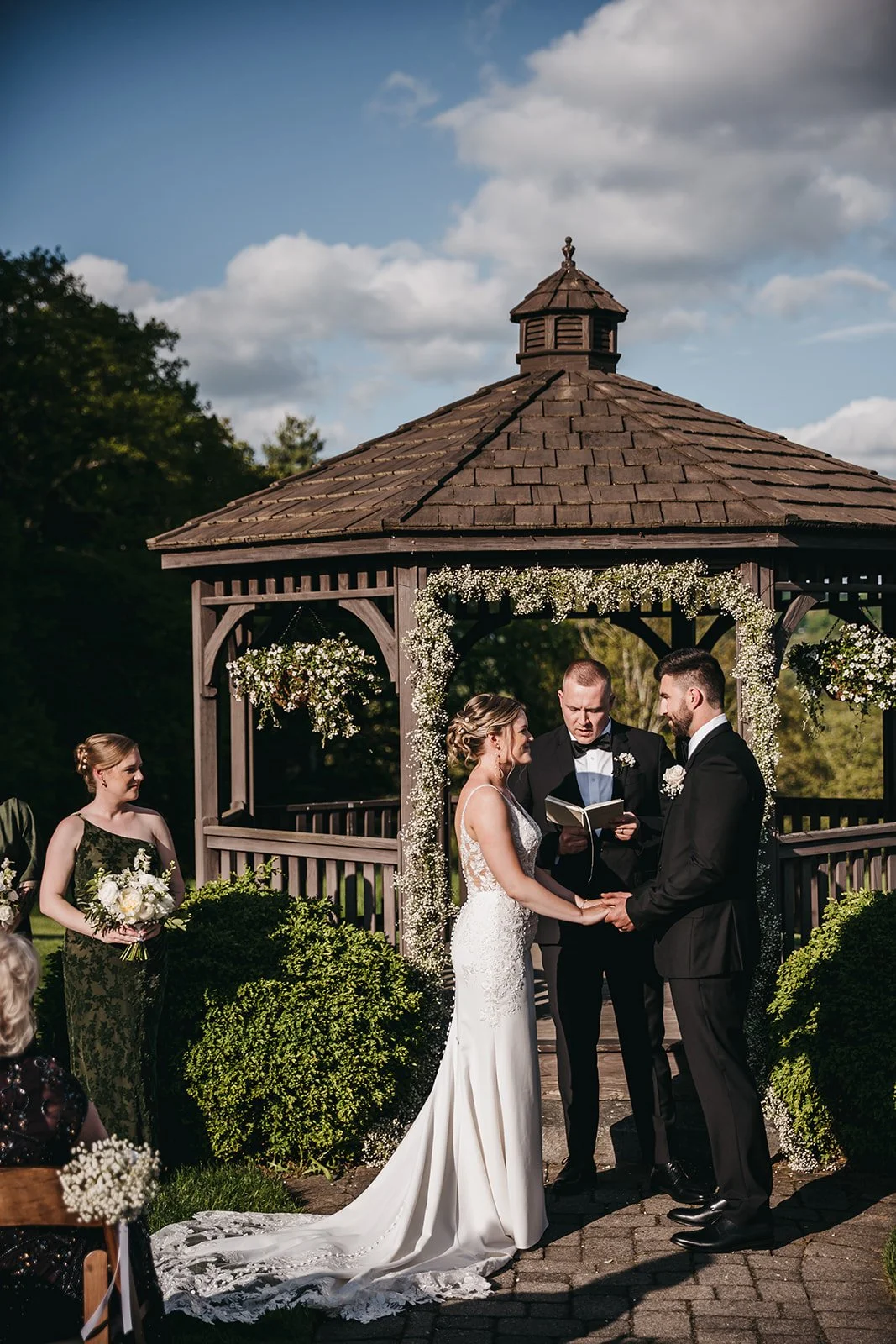 A wedding ceremony taking place outdoors under a wooden gazebo decorated with white flowers. The bride and groom are holding hands and exchanging vows, with an officiant reading from a book. Bridesmaids and guests are visible nearby.