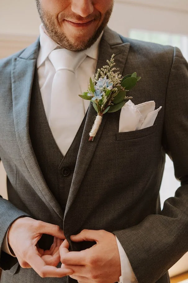 Close-up of a man in a gray suit with a boutonniere on his lapel and a white pocket square, adjusting his jacket.