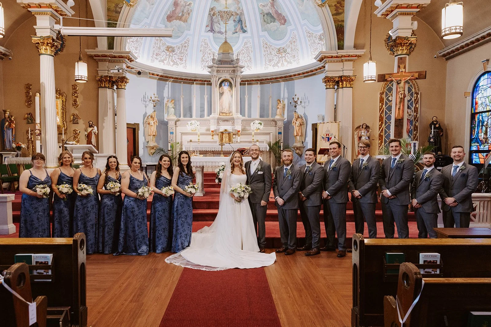 A wedding party stands inside a church with ornate walls, statues, and stained glass windows. The bride and groom are in the center, surrounded by bridesmaids in blue dresses on the left and groomsmen in gray suits on the right.