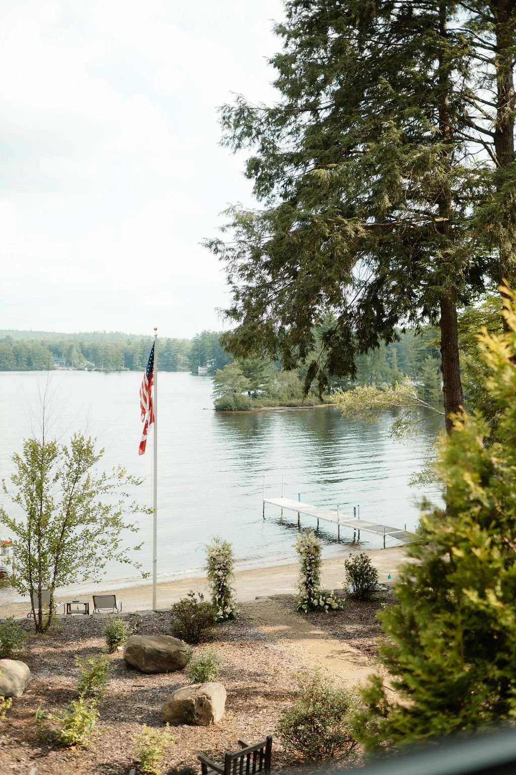 A lakeside scene with a flagpole flying the American flag, a dock extending into the water, and trees surrounding the area, taken from a porch or window.