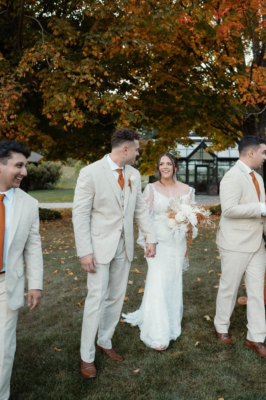 A bride in a white wedding dress holding a bouquet of flowers, walking hand in hand with the groom, surrounded by groomsmen dressed in beige suits with orange ties, outdoors during autumn with colorful trees in the background.
