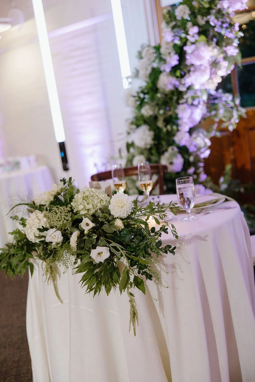 Elegant wedding reception table with white floral centerpiece, glasses of champagne and water, set against a backdrop of purple lit floral arrangement.