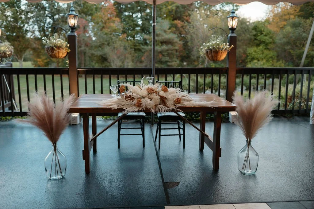 A decorated outdoor patio table with floral arrangements on a wooden table, surrounded by black chairs, with hanging flower baskets on wooden posts and lantern-style lights, overlooking a wooded area with fall foliage.