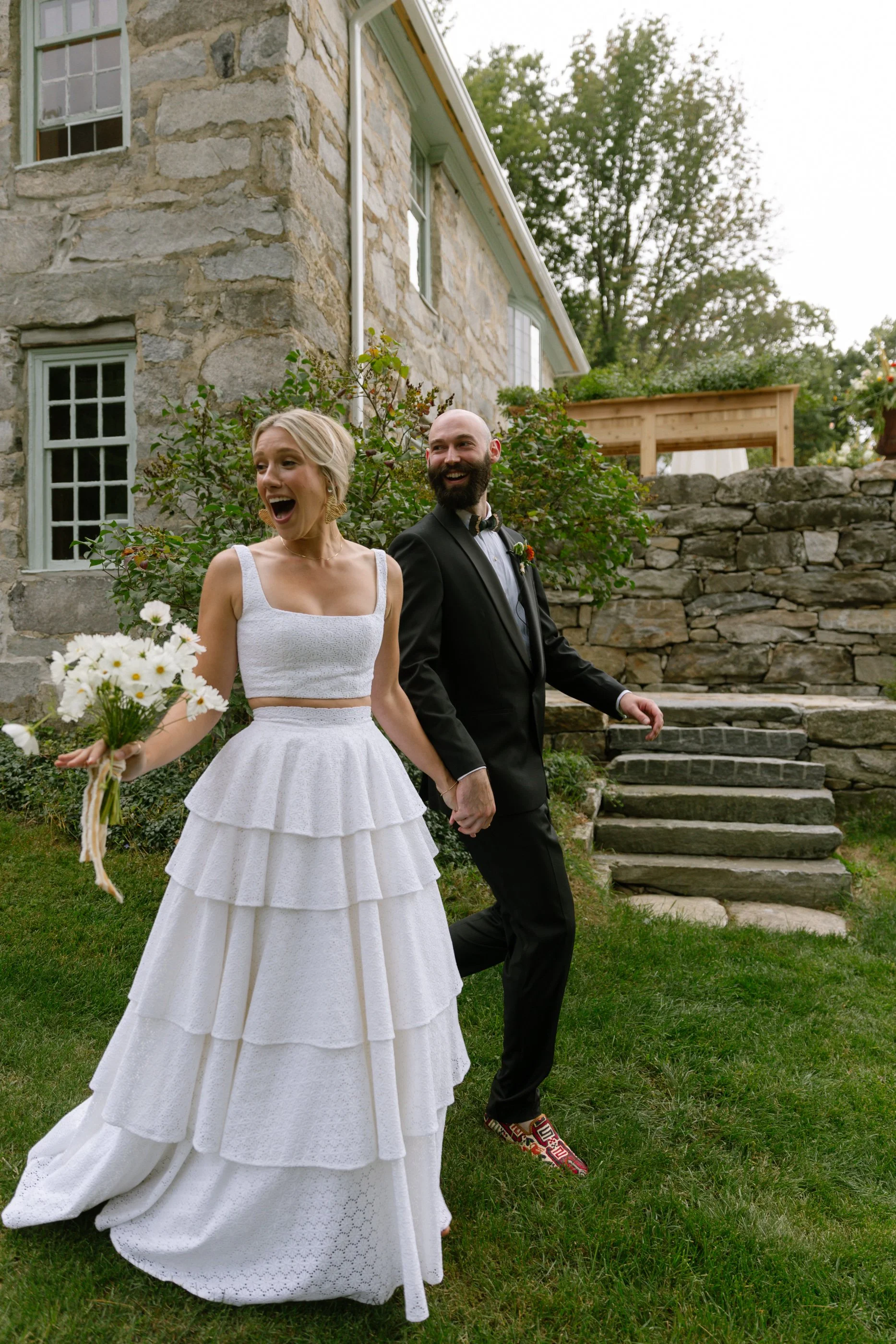 A bride in a white tiered dress holding a bouquet of daisies, and a groom in a black suit with a bow tie, walking hand in hand outside near a stone house with steps and greenery, smiling and celebrating.
