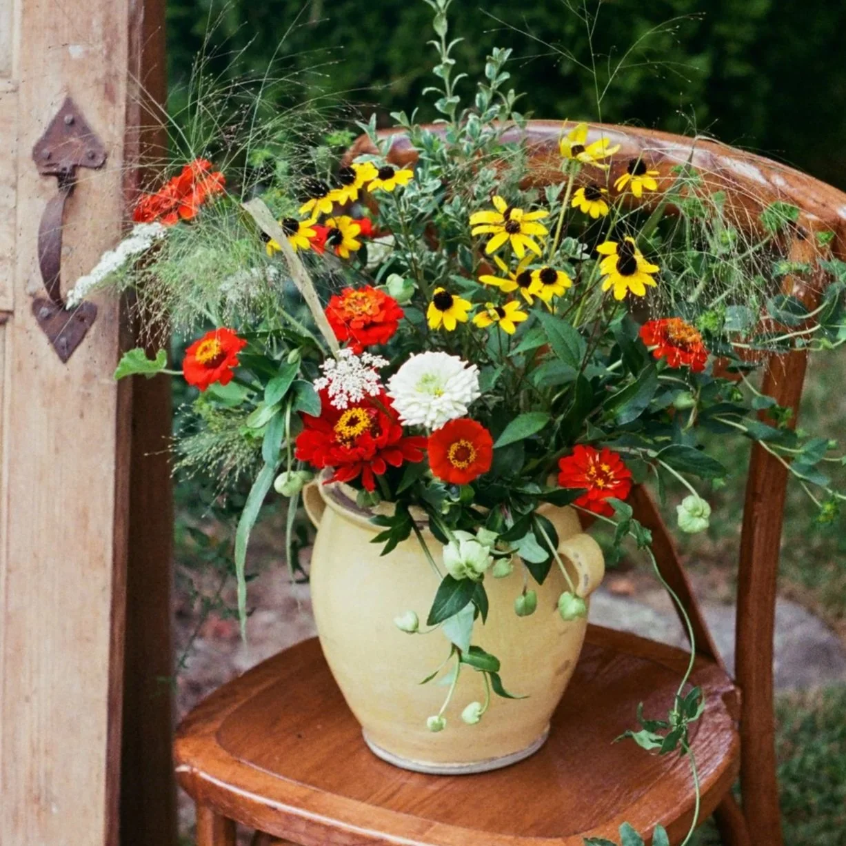 A ceramic yellow vase filled with a colorful bouquet of red, yellow, and white flowers, with green foliage and delicate grasses, placed on a wooden chair outdoors.