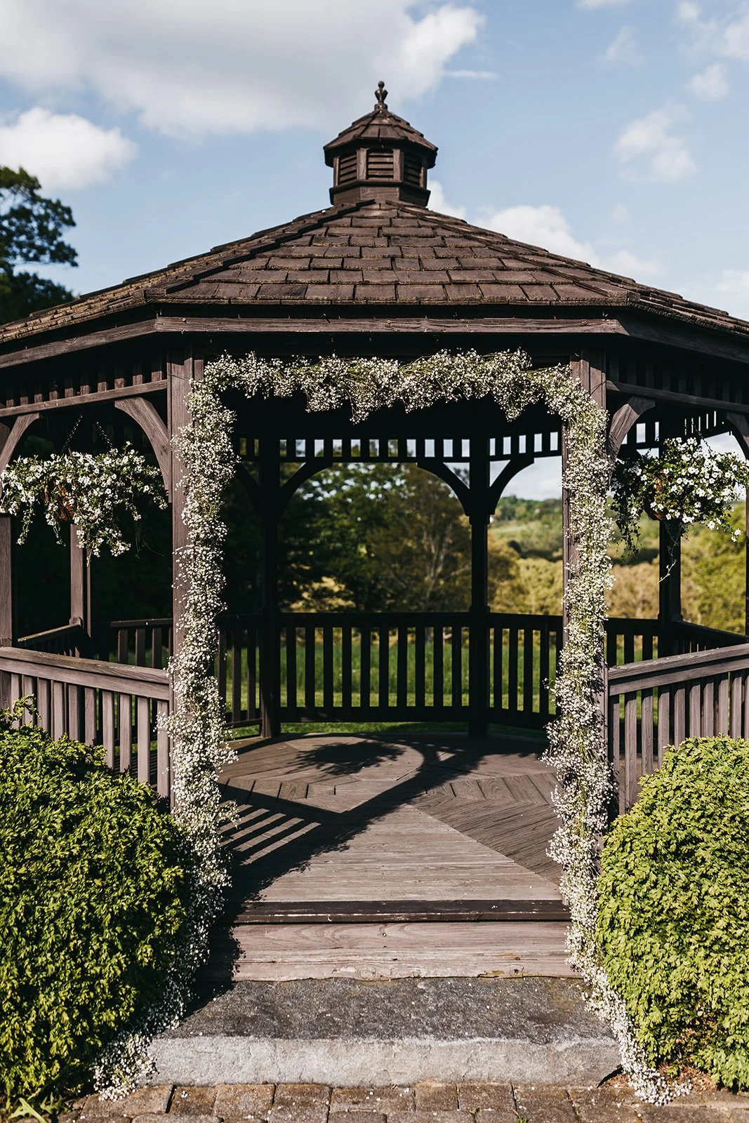 Wooden gazebo decorated with white flowers and greenery at the entrance, outdoors on a sunny day.