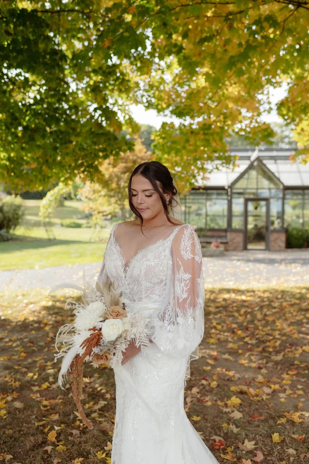 A bride in a white lace wedding dress holding a bouquet of white and beige flowers, standing outdoors under a tree with yellow and green leaves, with a greenhouse in the background.
