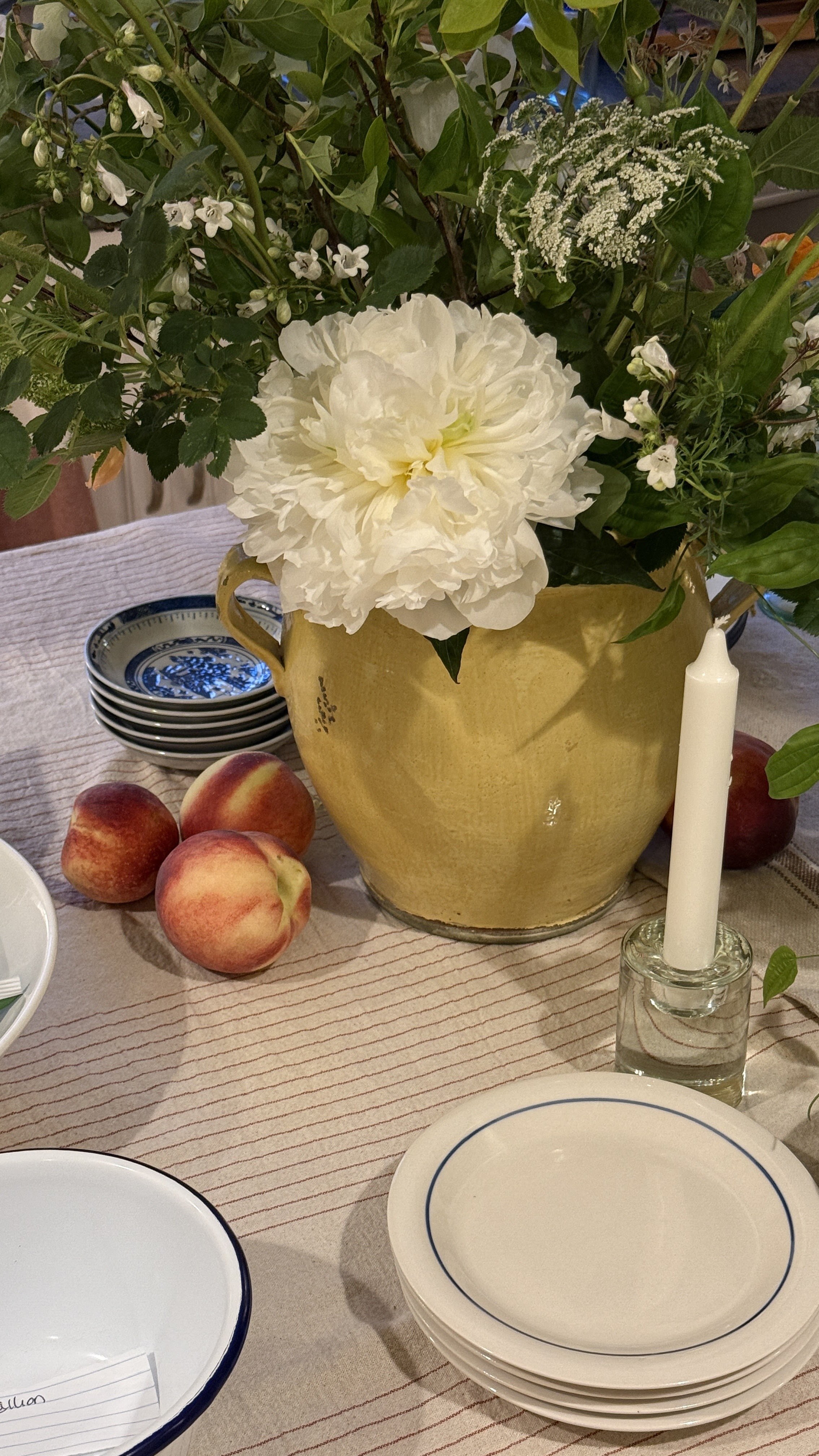 A yellow vase with white peonies and other greenery, surrounded by apples, some stacked blue and white dishes, a white candle in a glass holder, and a beige tablecloth with red stripes.
