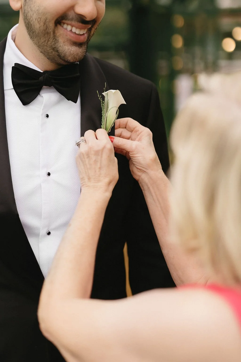 A woman pinning a white calla lily boutonniere on a man's black tuxedo at a wedding.