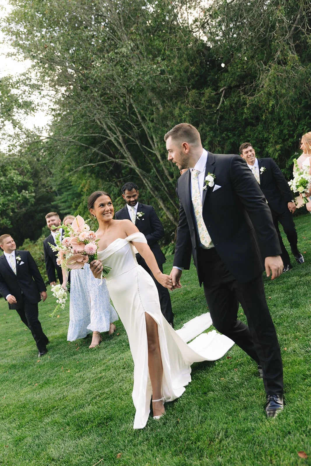 A bride and groom holding hands and walking on grass outdoors, surrounded by wedding guests. The bride is wearing a white gown and holding a bouquet, smiling at the groom. The groom is in a black suit and tie, looking at the bride. Guests are dressed