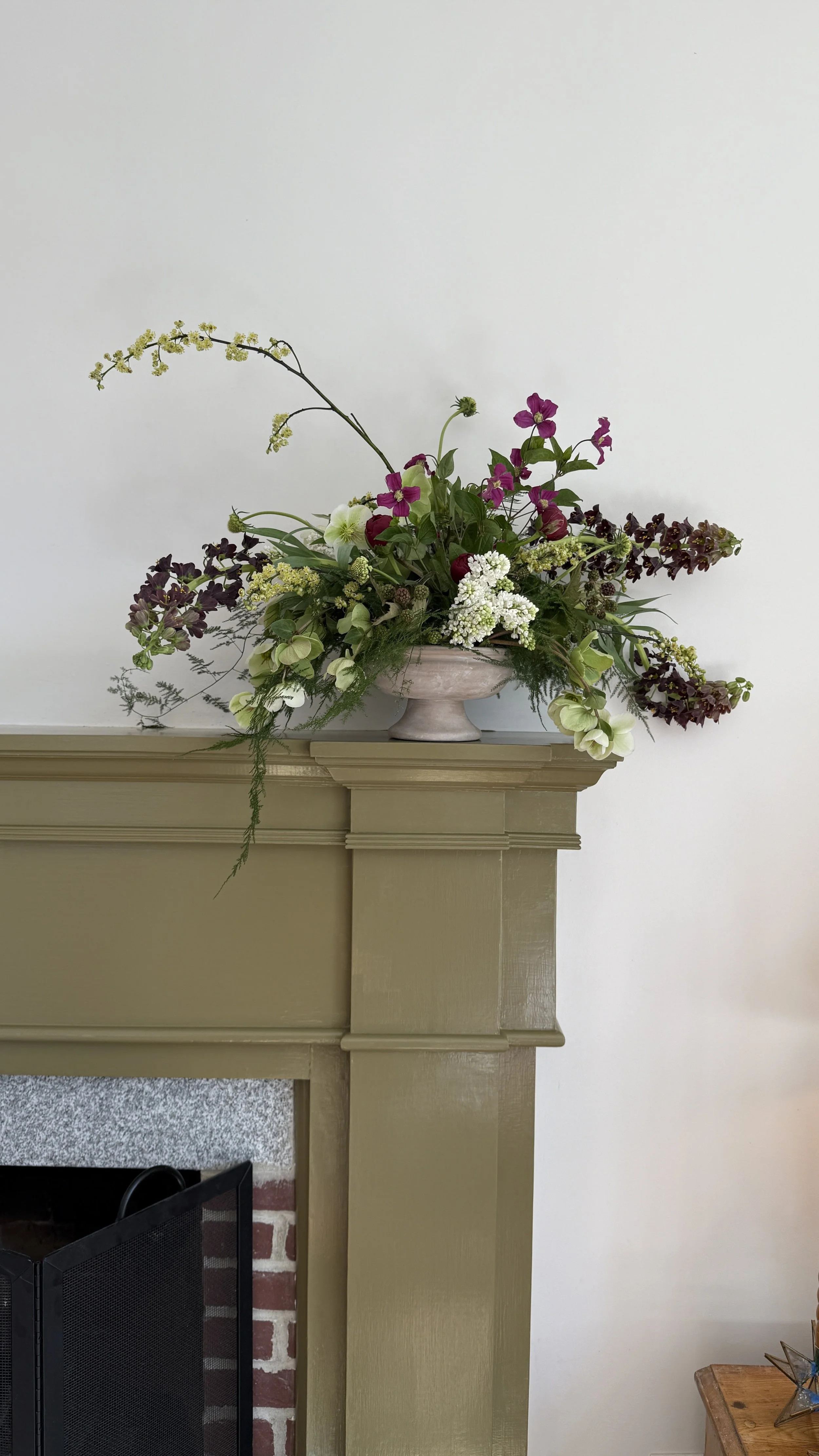 A floral arrangement with various flowers, including white, purple, and pink blooms, placed in a decorative white vase on top of a beige fireplace mantel.