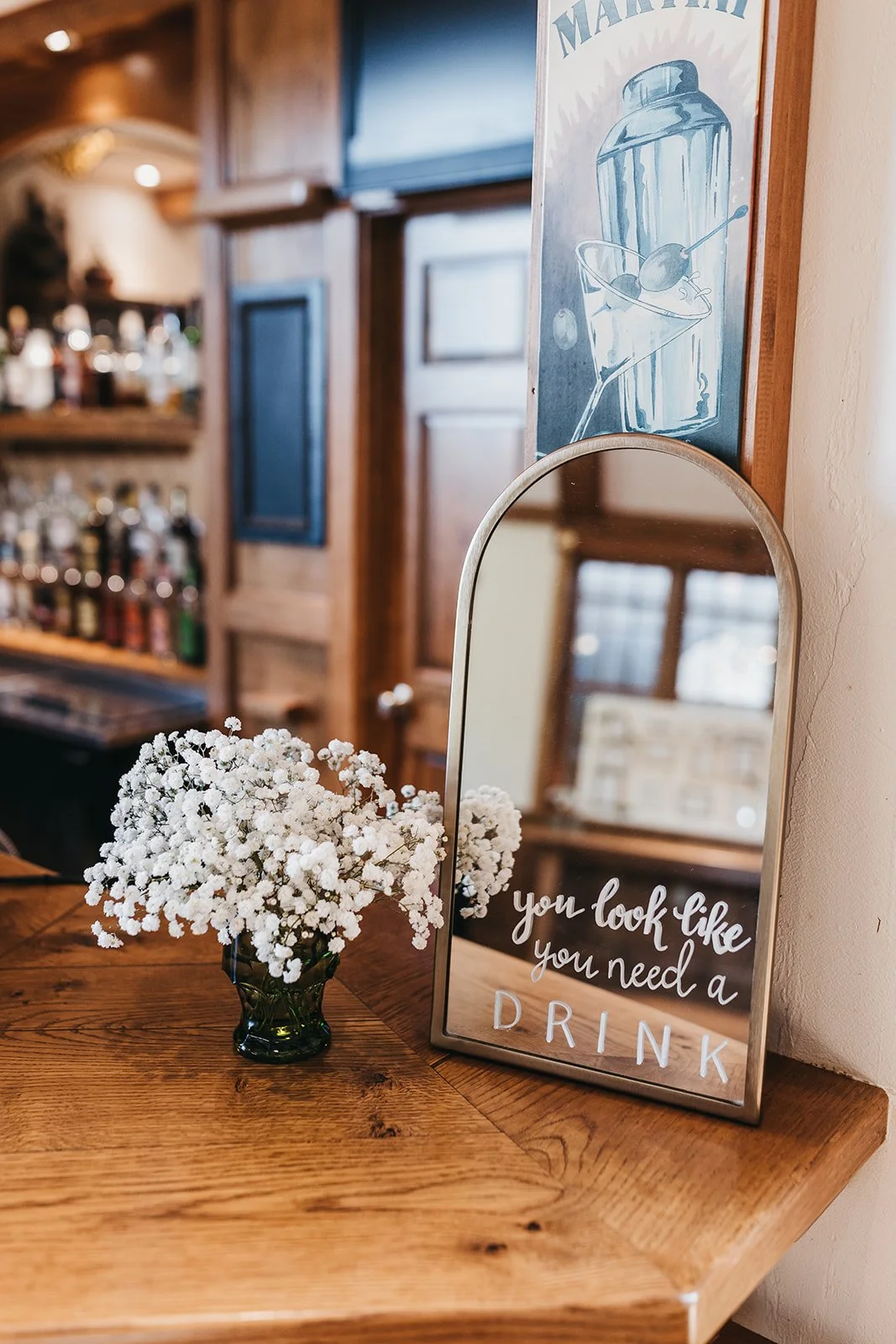A mirror with the text 'you look like you need a DRINK' on it, sitting on a wooden table next to a small bouquet of white baby's breath flowers in a dark glass vase. A wood-paneled bar with bottles is visible in the background.
