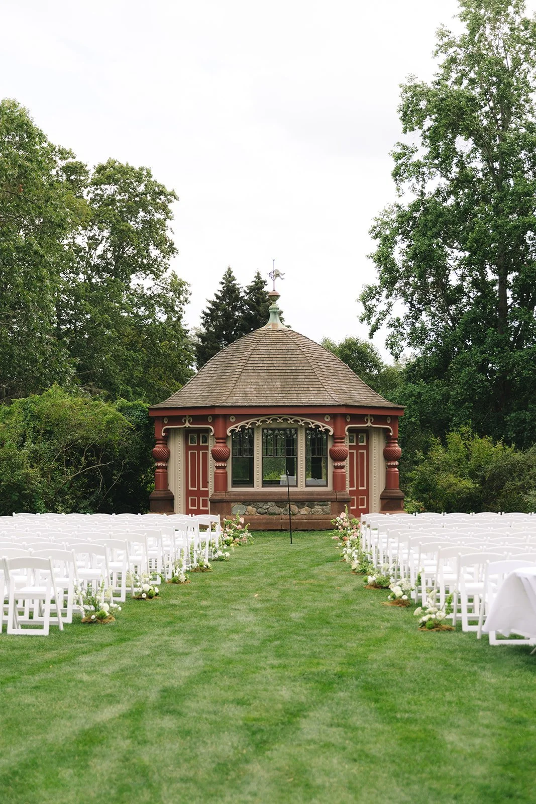 Outdoor wedding ceremony setup with rows of white chairs on either side of a grassy aisle, leading to a small ornate wooden pavilion surrounded by greenery.