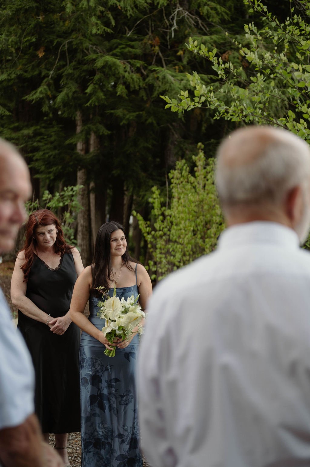A woman in a blue dress holding a bouquet of white flowers at an outdoor event, with other people and green trees in the background.