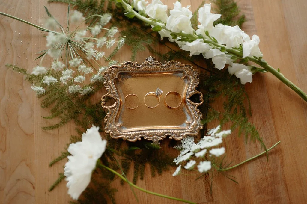 A vintage gold ring box with three rings inside, surrounded by white flowers and greenery on a wooden surface.