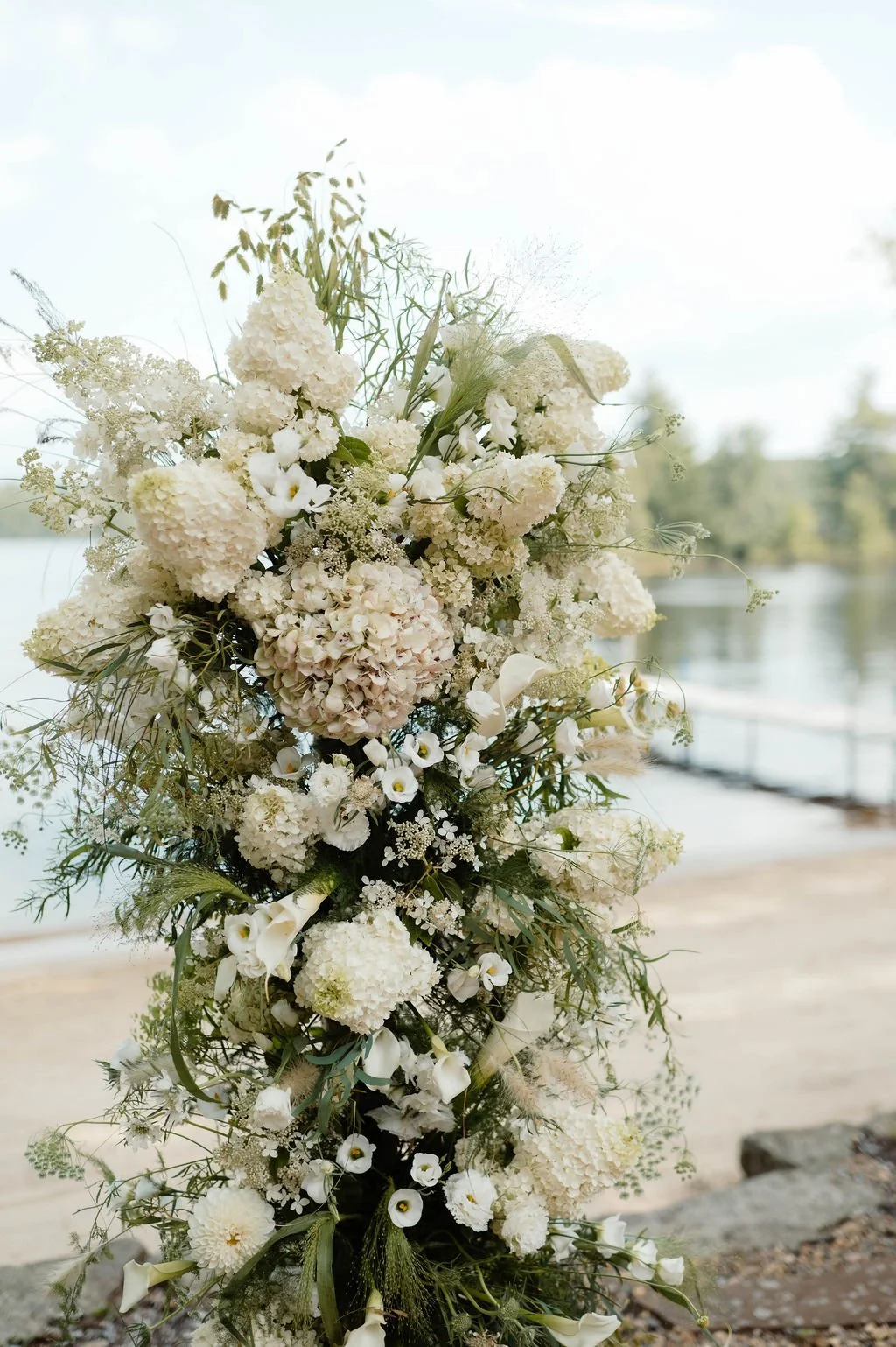A large arrangement of white and cream-colored flowers near a lake or river, with blurred trees and water in the background.