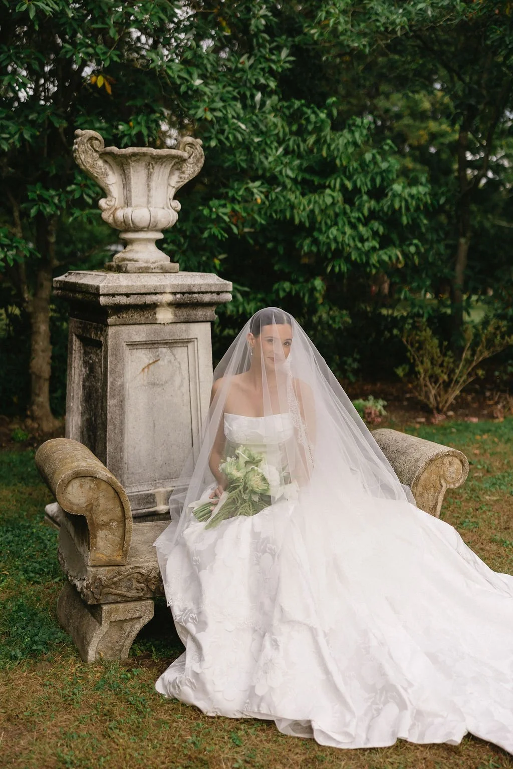 A bride in a white wedding dress and veil sitting on a stone bench outdoors, holding a bouquet of white flowers, with a large stone urn and lush green trees in the background.