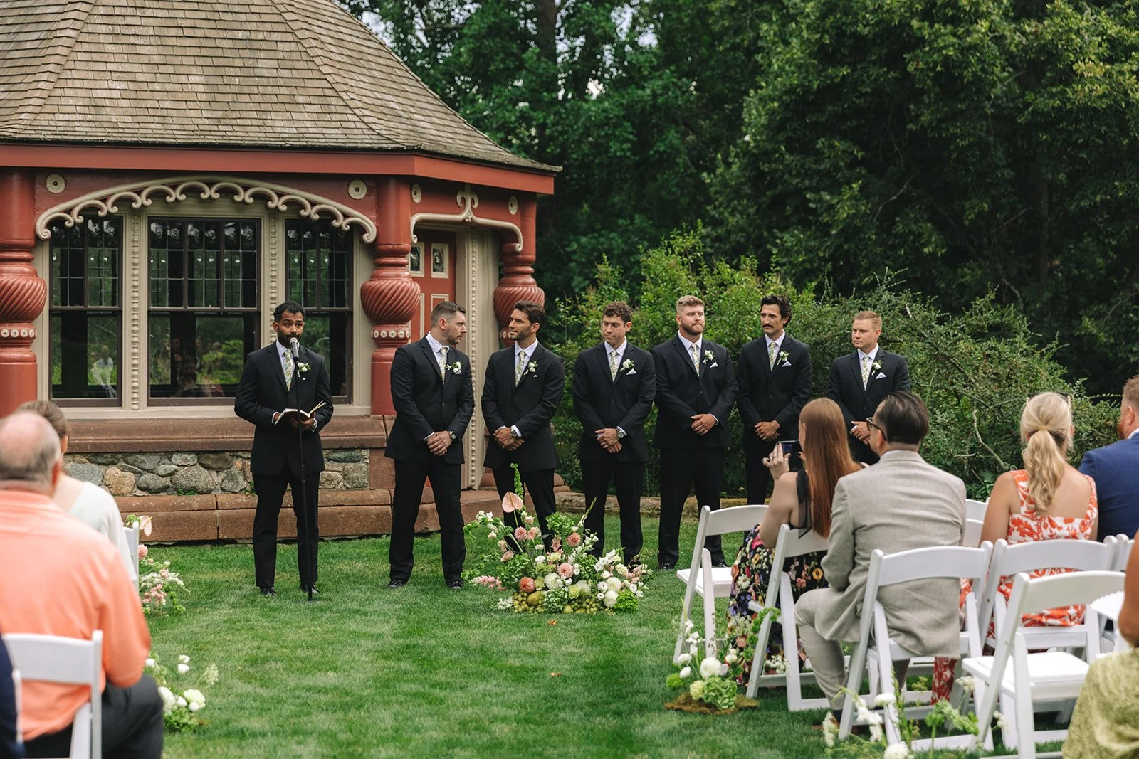 Groom and groomsmen standing in a line at an outdoor wedding ceremony, with guests seated in front of them, on a lush lawn with floral arrangements, in front of a decorative building with large windows and greenery.