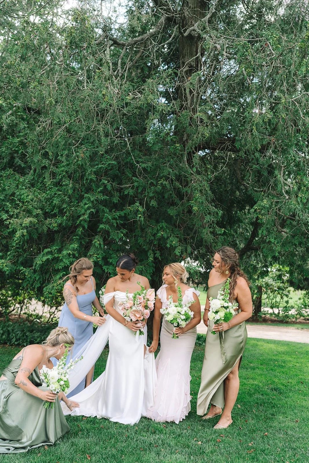 A bride in a white wedding dress with pink flowers in her bouquet stands with five bridesmaids in pastel-colored dresses holding white floral bouquets, gathered outdoors under a large leafy tree.