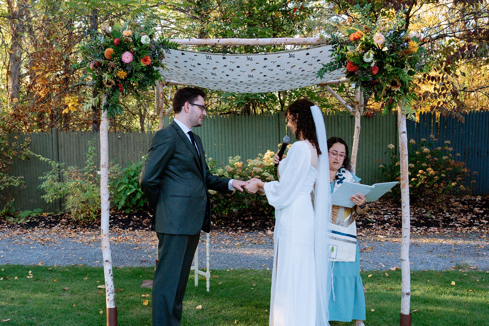 A couple gets married outdoors under a decorated canopy, with an officiant reading from a book and holding a microphone. The groom wears a dark suit and glasses, while the bride wears a white dress and veil. The setting has trees and a green fence in
