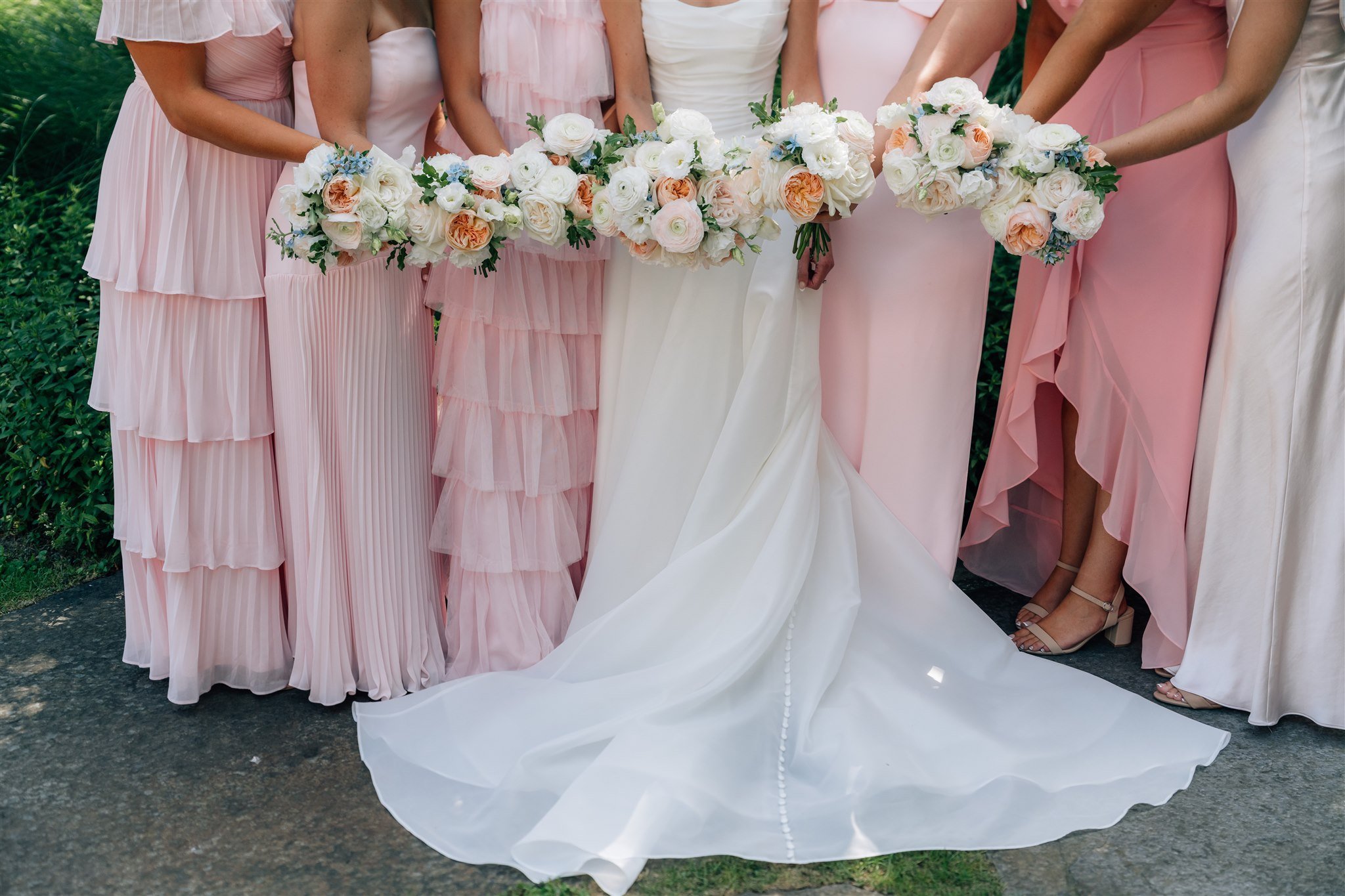 A group of women, likely bridesmaids and a bride, are standing outdoors wearing pastel pink and white dresses, holding bouquets of white and blush flowers.