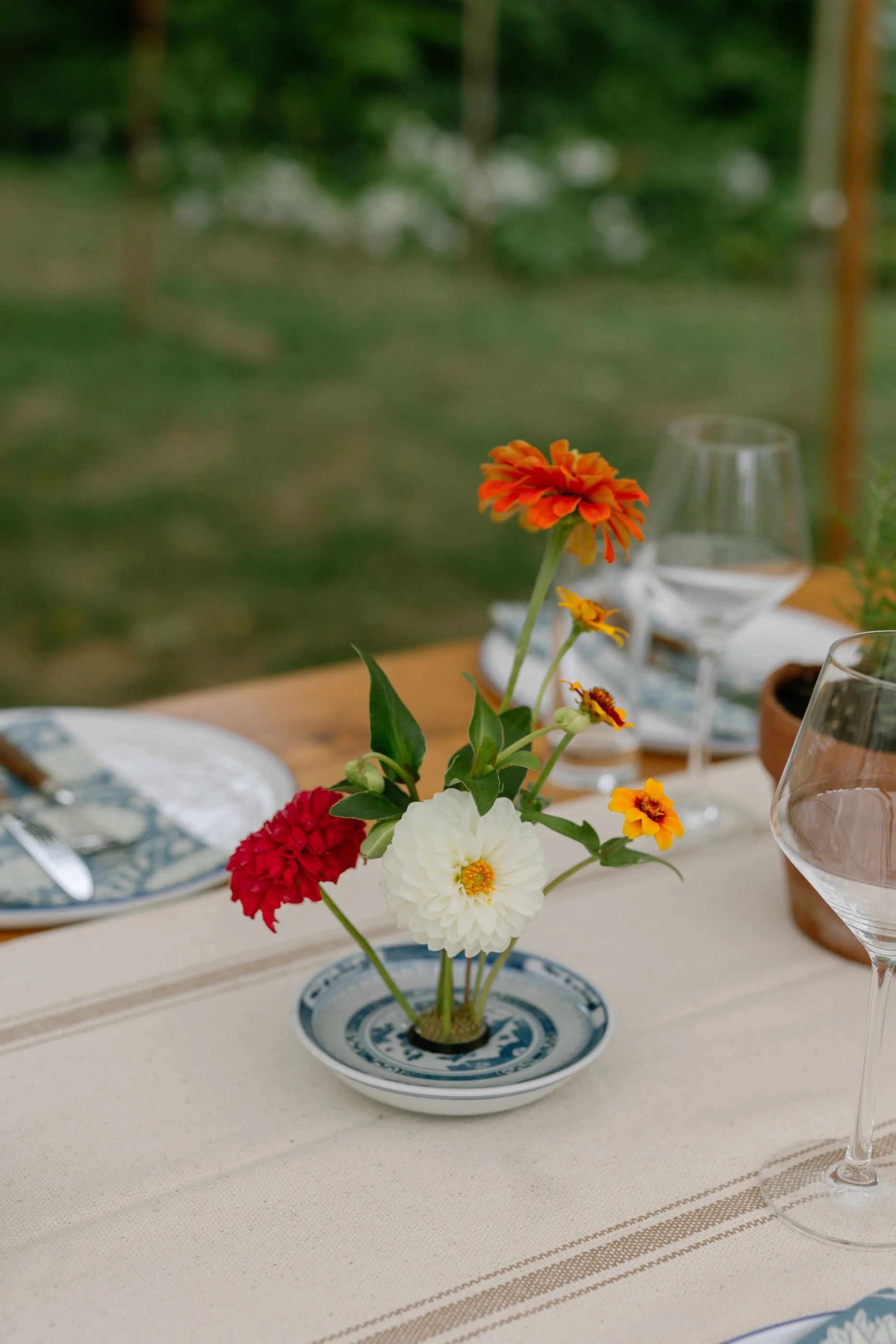 Colorful flower arrangement with orange, red, white, and yellow flowers on a dining table set outdoors.