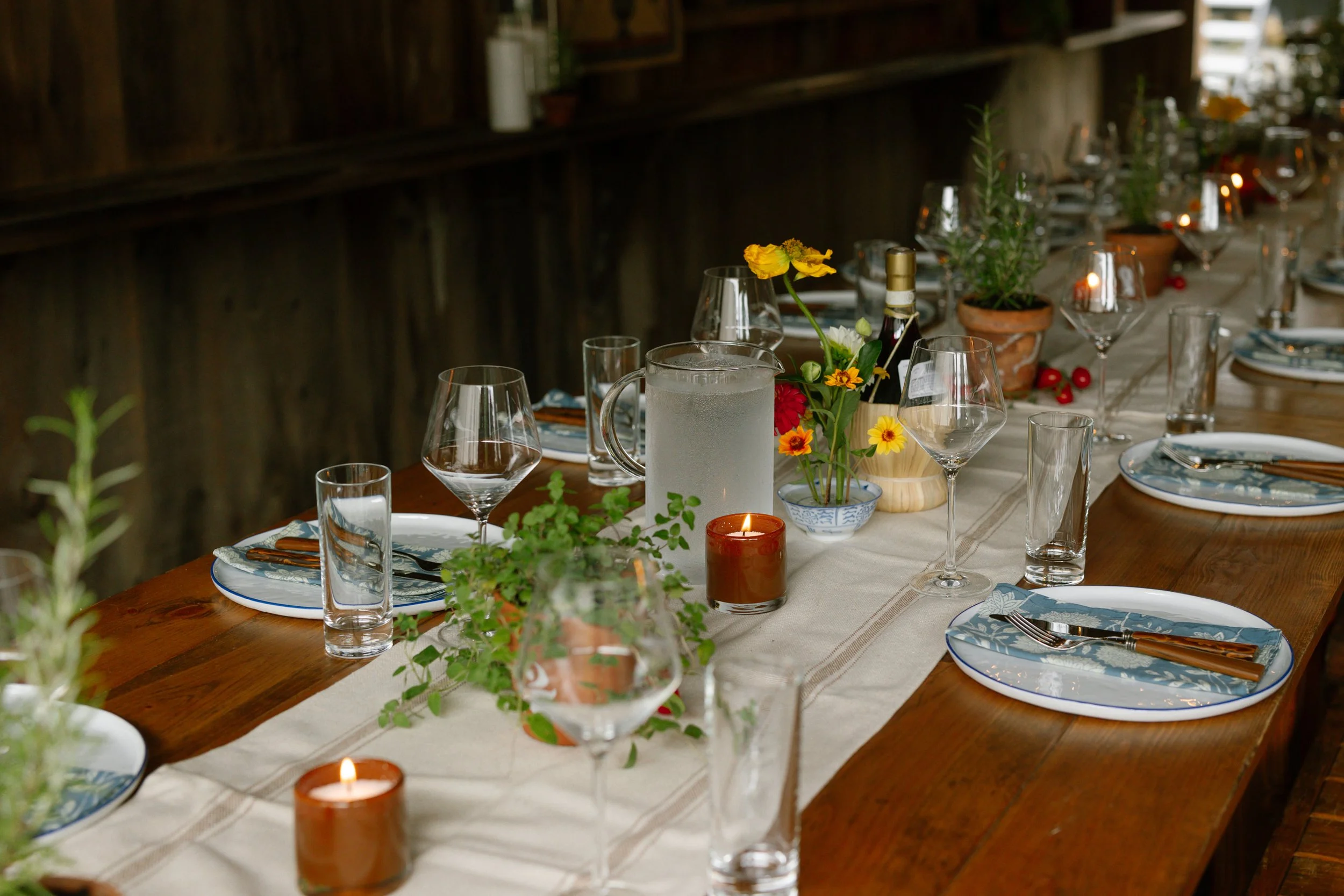 A decorated dining table with plates, cutlery, wine glasses, candles, potted plants, a pitcher of water, and a vase with flowers, set in a rustic wooden room.