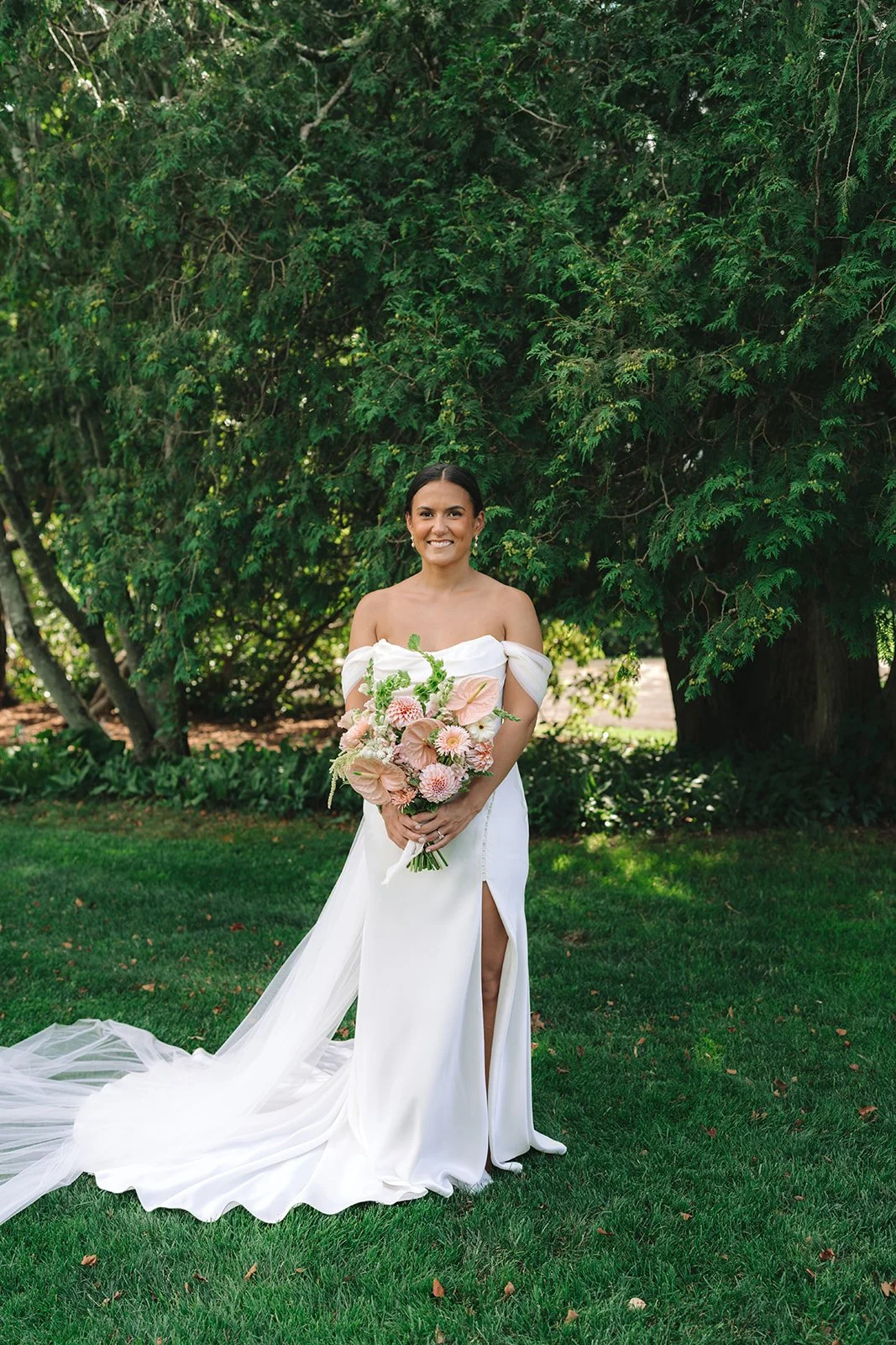 Bride standing outdoors in front of green trees, holding a bouquet of pink and white flowers, wearing a white off-shoulder wedding gown with a thigh-high slit.