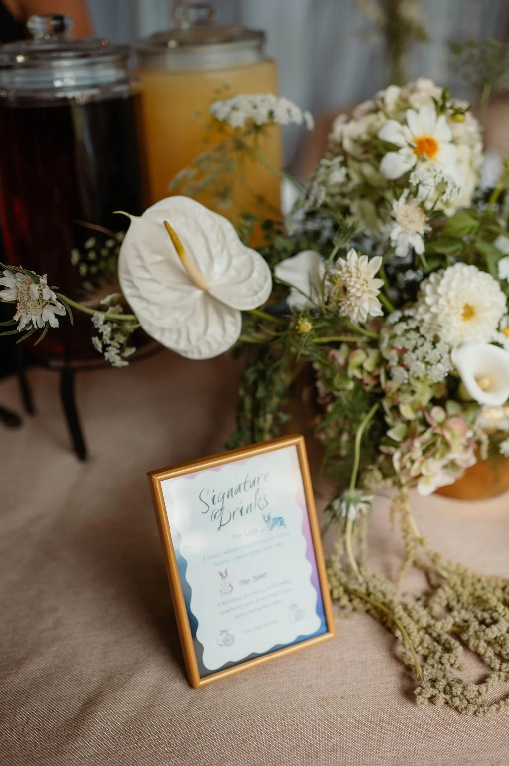 A table display with a framed sign for signature drinks, surrounded by white flowers and containers with drinks in the background.