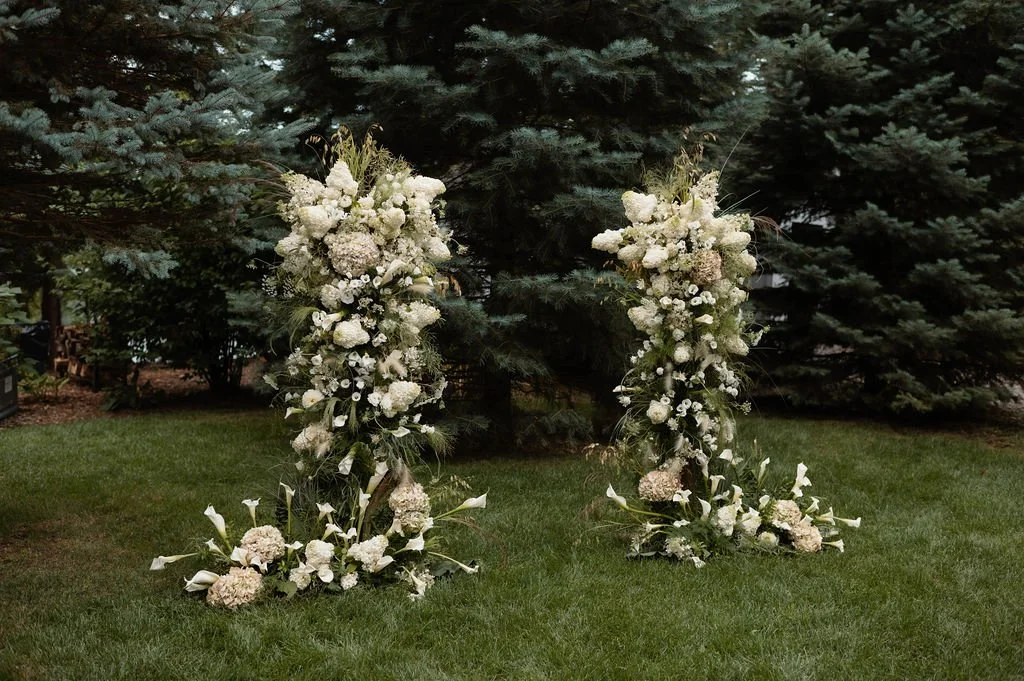 Two floral archways decorated with white flowers and greenery on a grassy outdoor area with trees in the background.