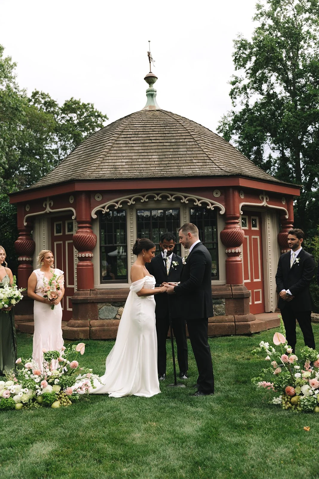 A wedding ceremony taking place outdoors in front of a small, ornate red and beige pavilion with trees in the background. The bride and groom are holding hands and exchanging vows, with bridesmaids and groomsmen standing nearby, along with floral arr
