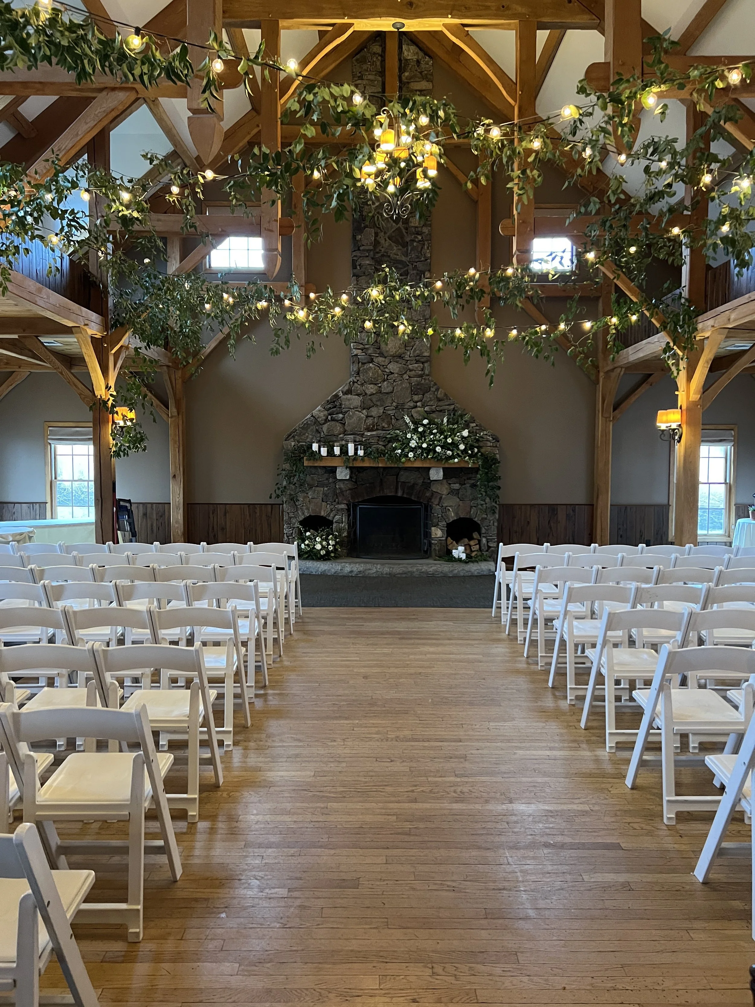 An indoor wedding venue with wooden beams, string lights, and a stone fireplace decorated with flowers and greenery. White chairs are arranged facing the fireplace on a wooden floor.