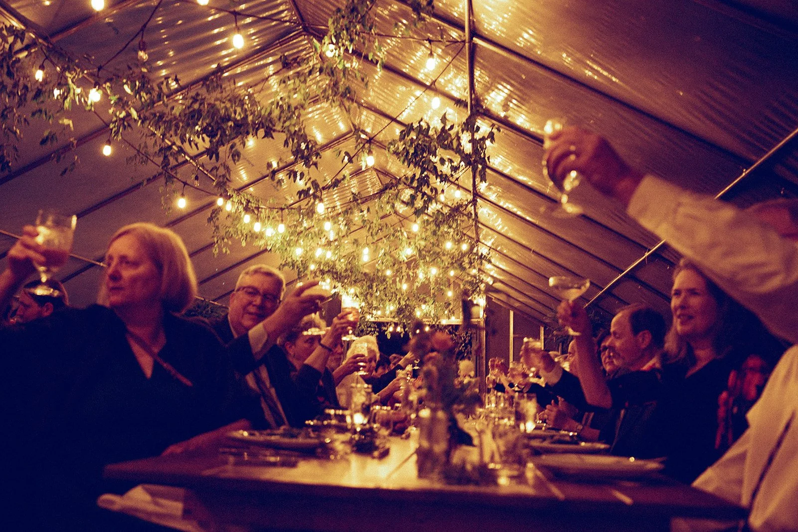 People sitting at a long dining table inside a tent, holding drinks, under string lights and greenery decorations.