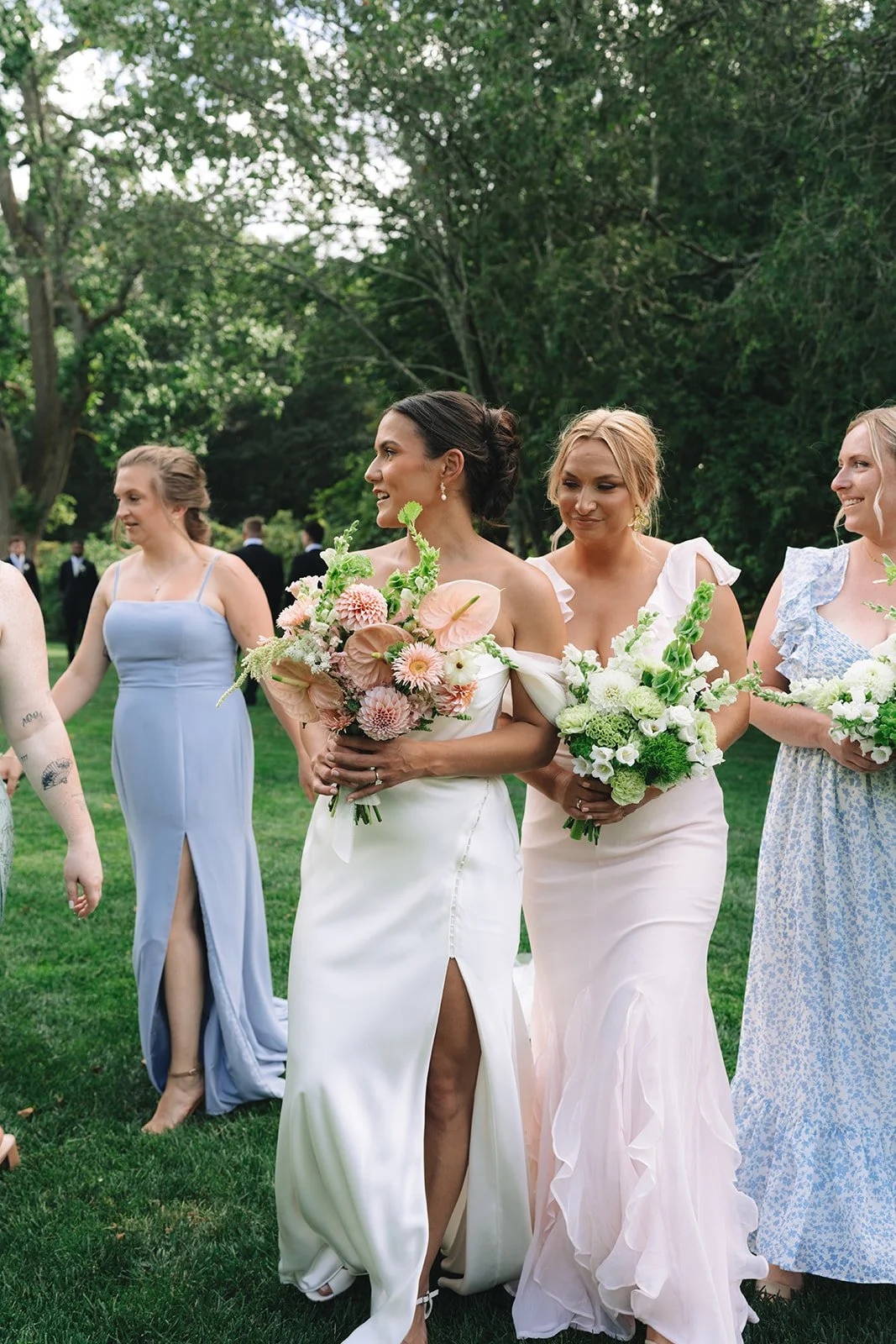 A group of women at a wedding, dressed in pastel gowns, holding bouquets of flowers outdoors on green grass with trees in the background.