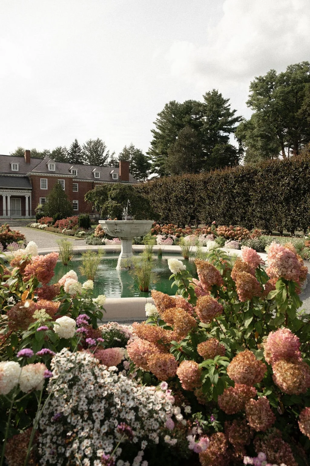 A landscaped garden with a fountain at the center, surrounded by colorful flowers and lush green trees, with a large brick building in the background and a cloudy sky overhead.