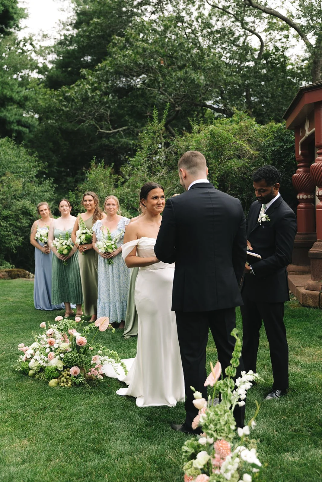 Bride and groom exchanging vows outdoors, surrounded by bridesmaids holding bouquets, during a wedding ceremony in a garden with lush greenery and floral arrangements.