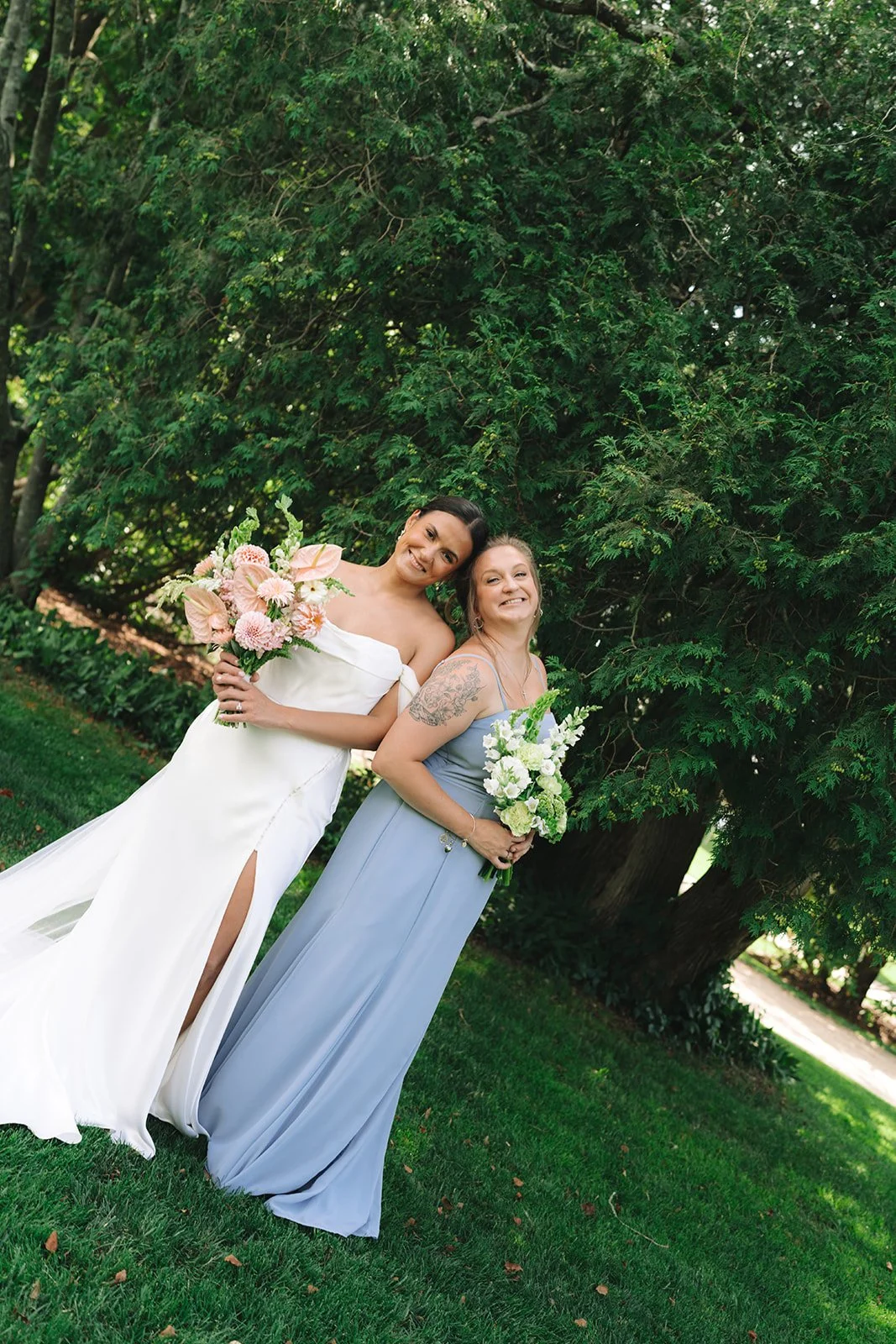 Two women standing on grass in front of a large tree, smiling and holding bouquets of flowers, one in a white dress and the other in a light blue dress.