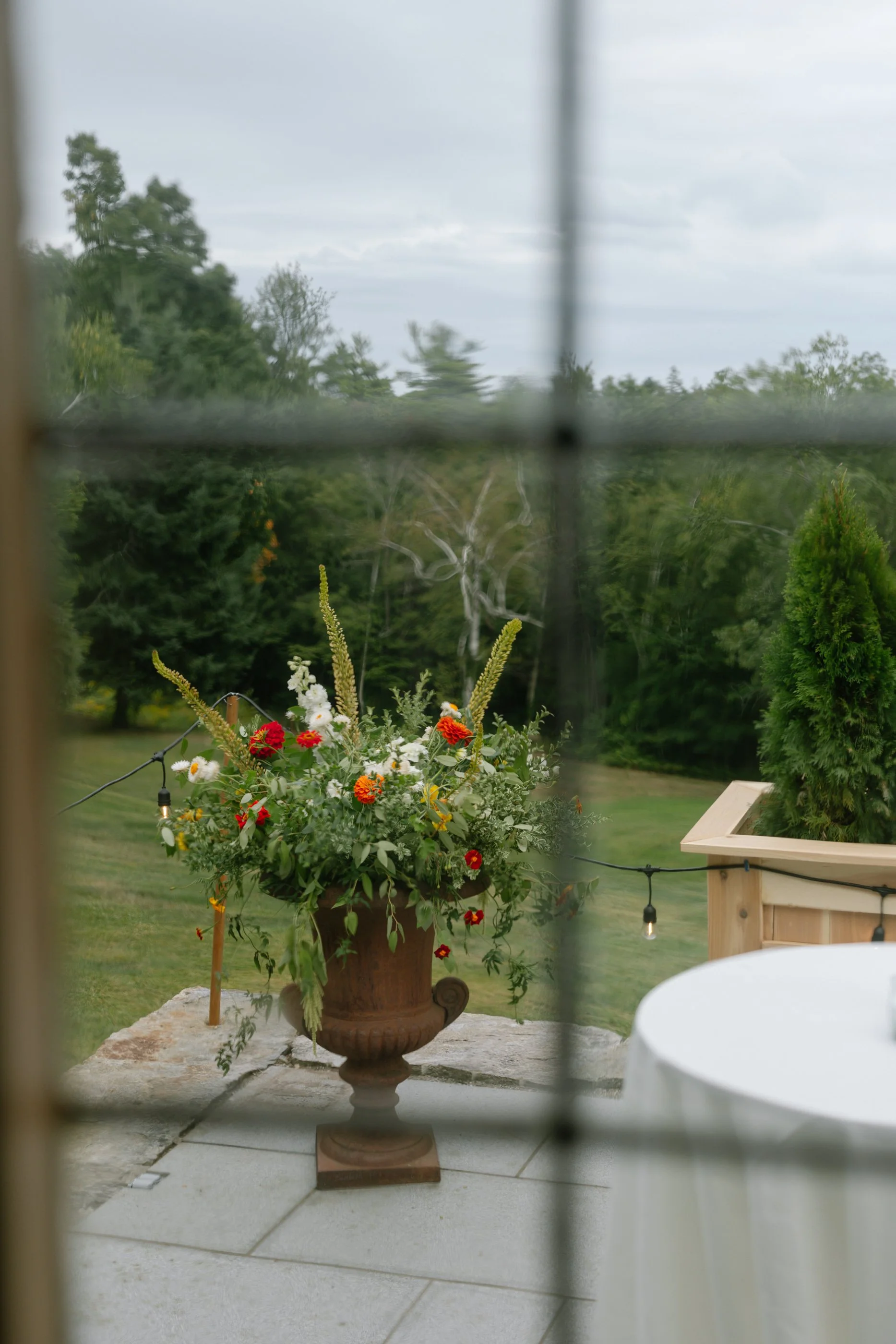 View through a window showing a patio with a large flower arrangement in a classical urn, greenery, trees, and overcast sky in the background.