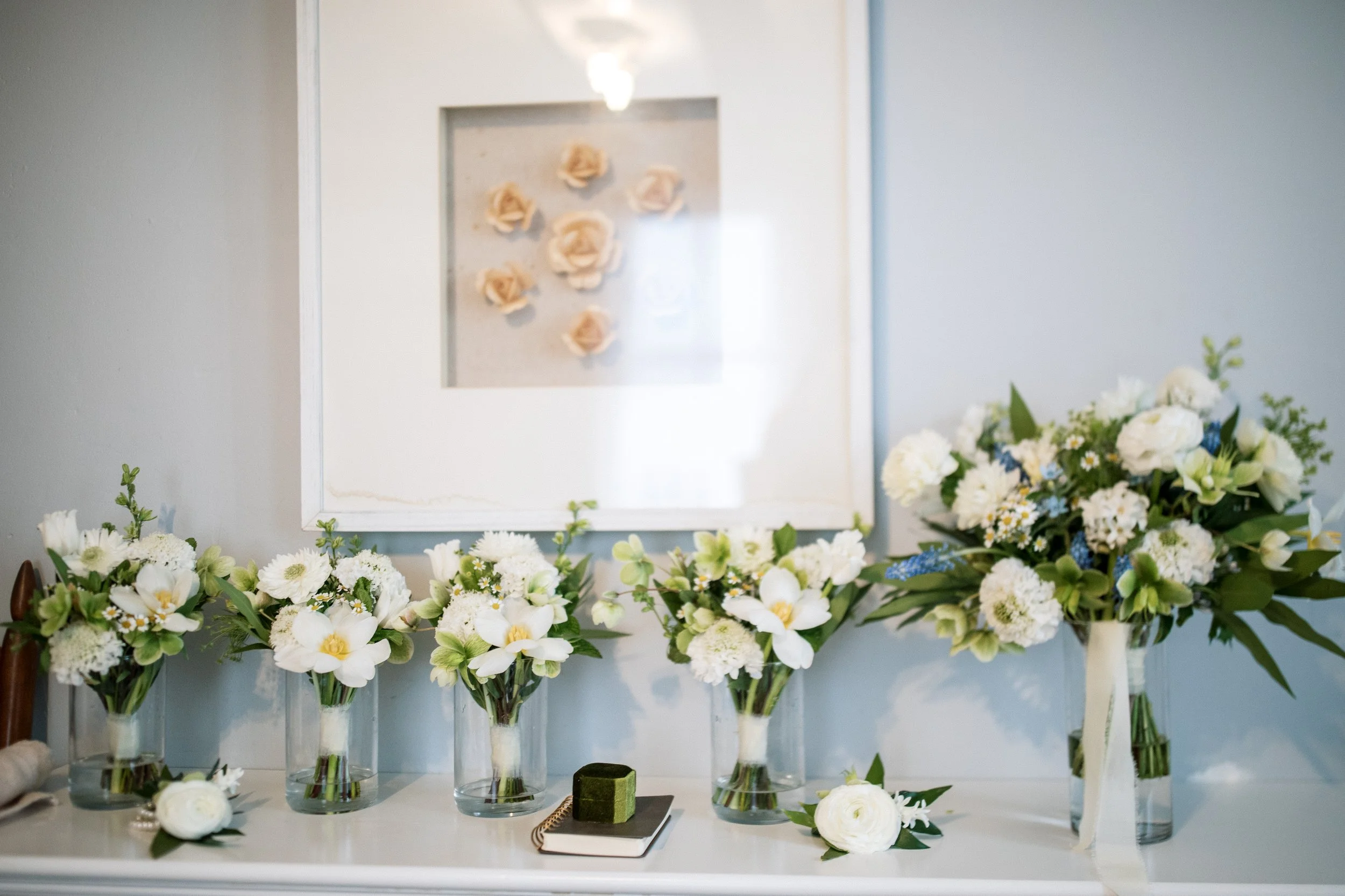 Several white flower bouquets in vases arranged on a white table in front of a mirror with beige roses inside.