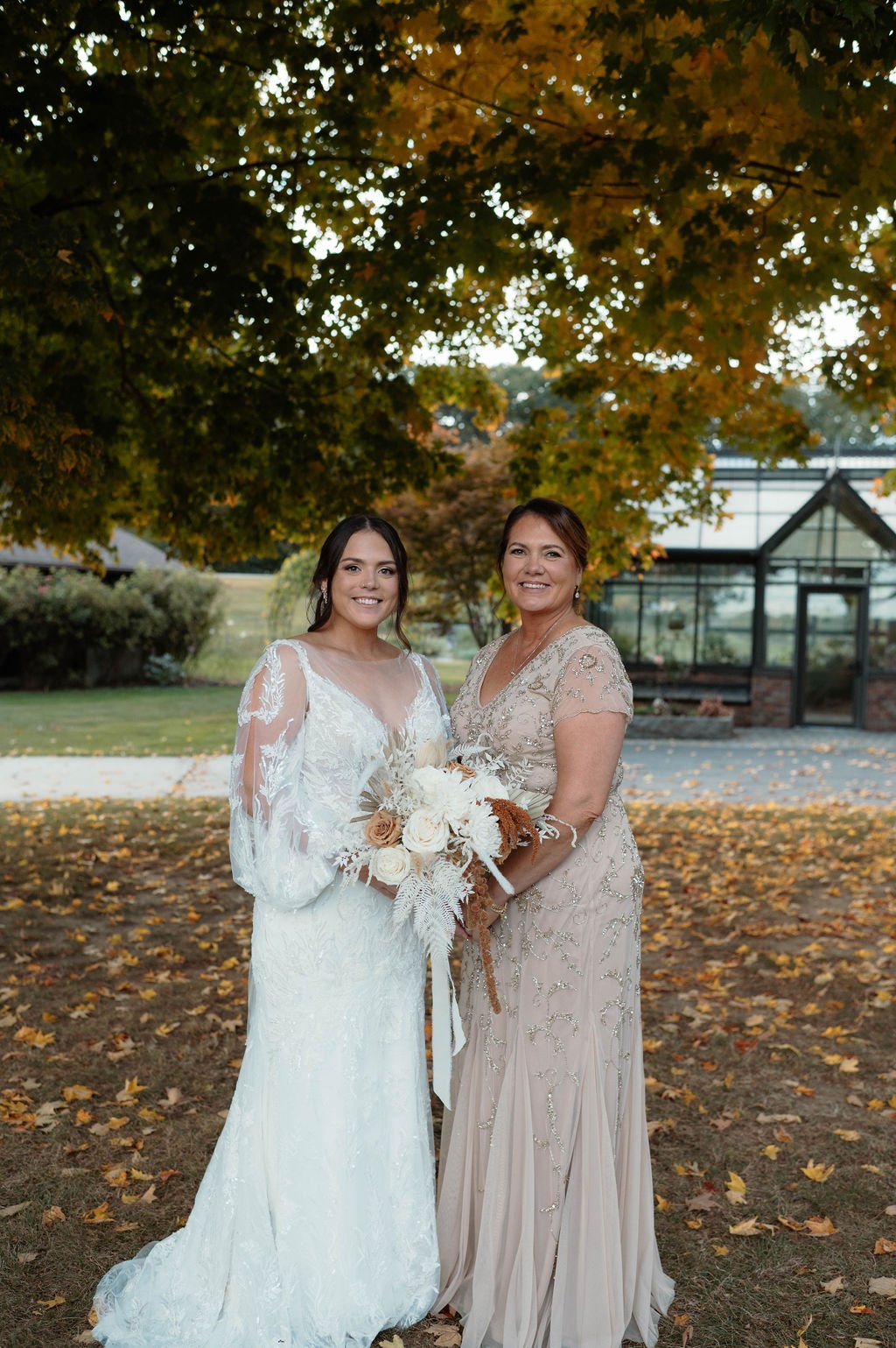 A bride and a woman, possibly her mother, standing outdoors under a large tree with autumn leaves, both smiling, with the bride holding a bouquet of flowers.