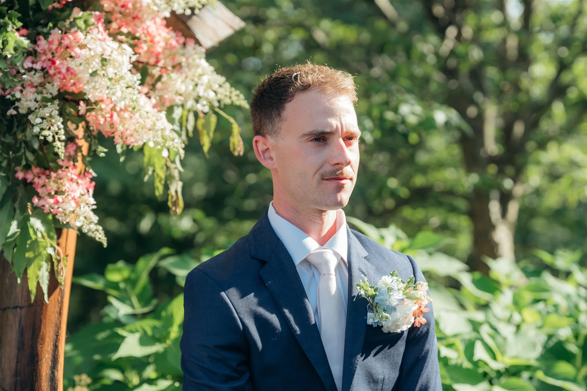 A groom standing outdoors in a garden during daytime, wearing a navy blue suit with a white tie and boutonniere, looking thoughtfully to the side, with pink and white flowers on his lapel and in the background.