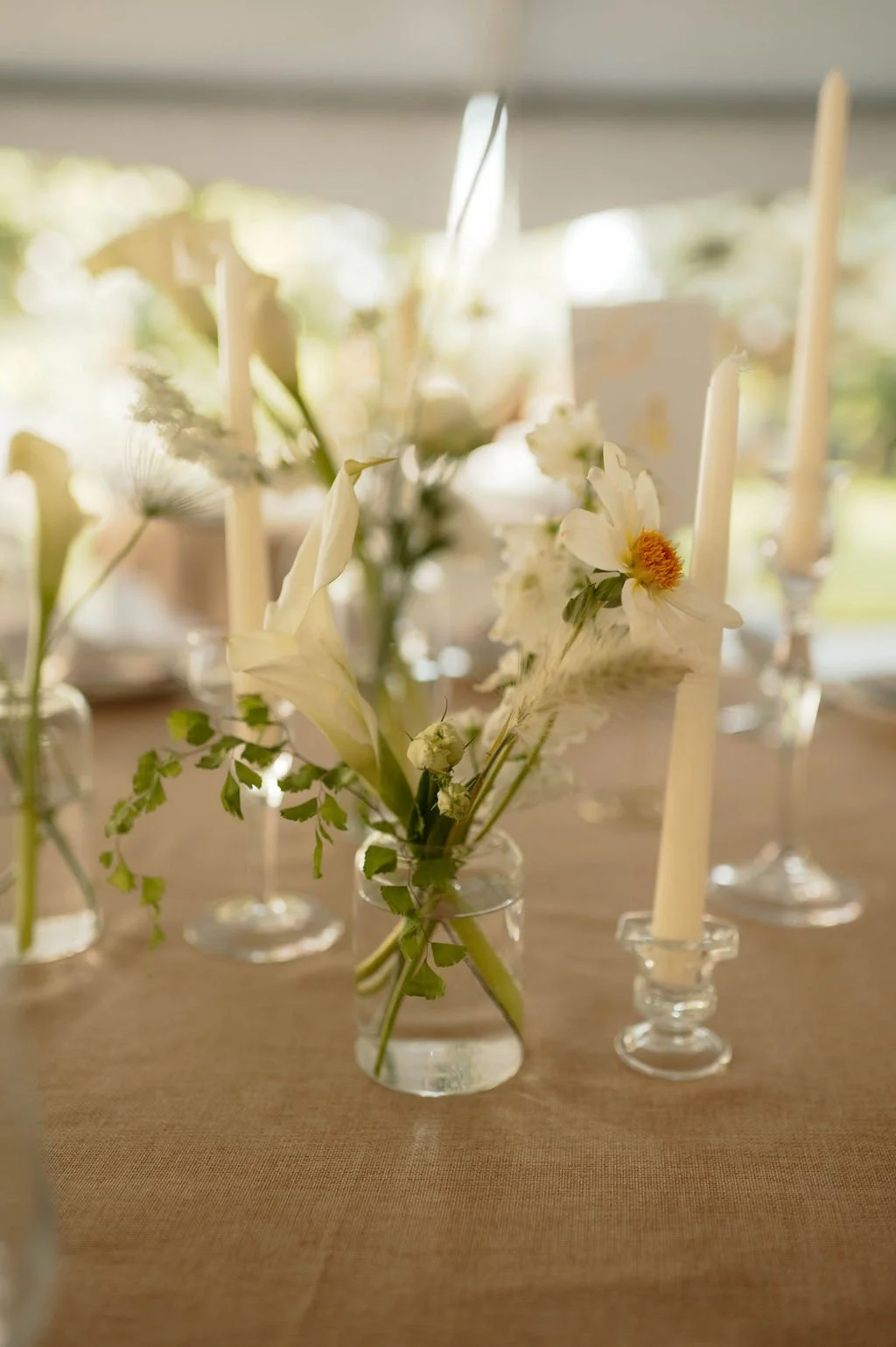Floral centerpiece with white flowers and greenery in a small glass vase, set on a wooden table with tall white candles in candleholders, in a softly lit setting.