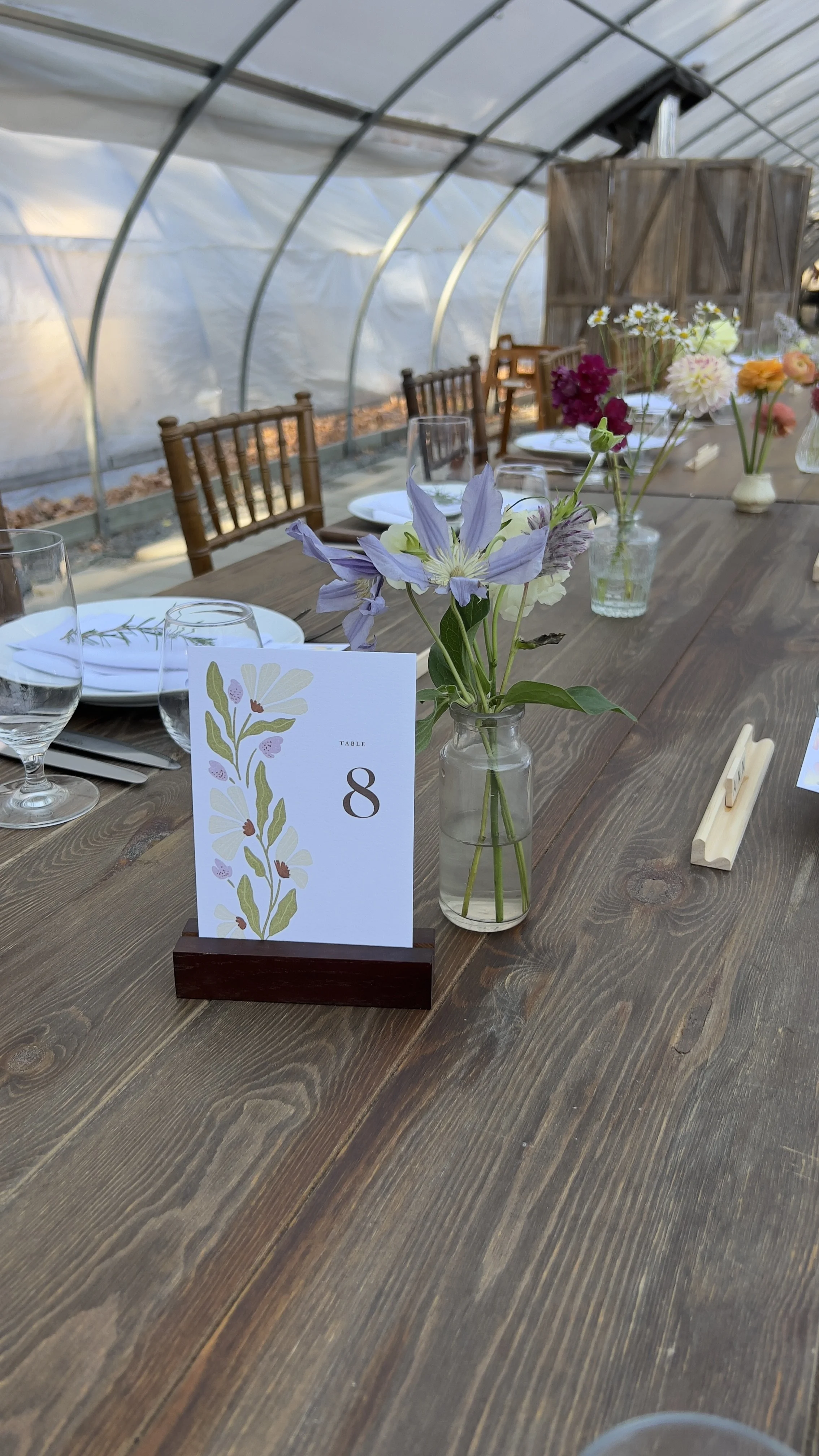 A decorated long wooden table with a table number card for table 8, and vases of fresh flowers, set inside a greenhouse with chairs around.