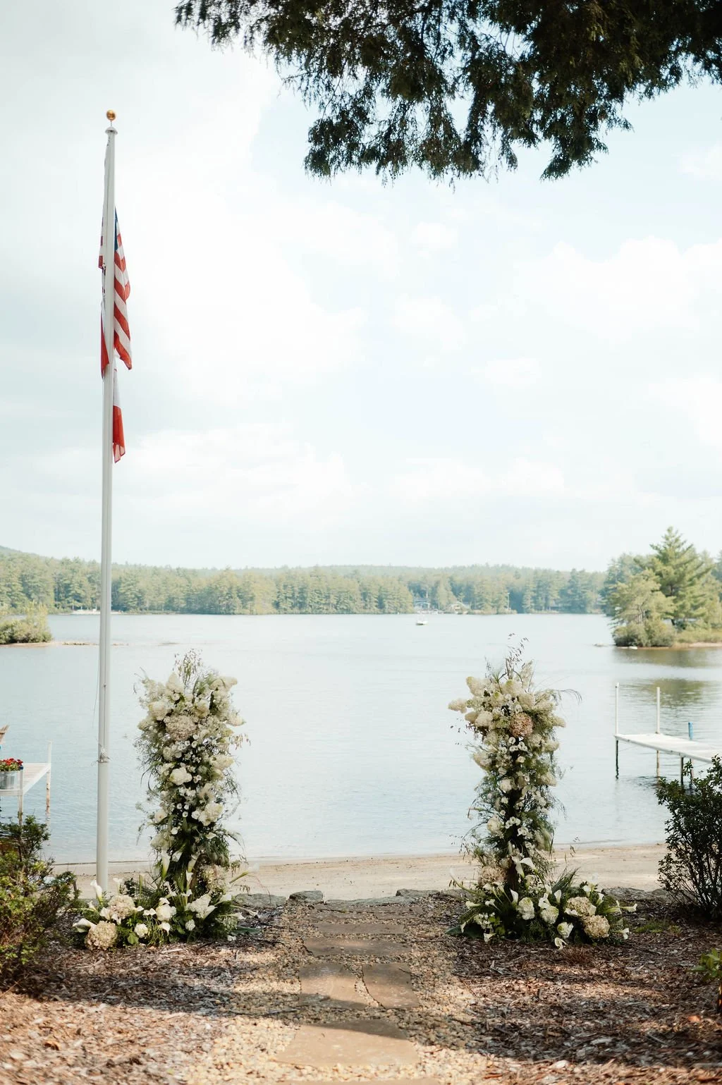 Wedding altar setup by a lakeside with floral arrangements, an American flag, and three steps leading to the water, surrounded by trees and cloudy sky.