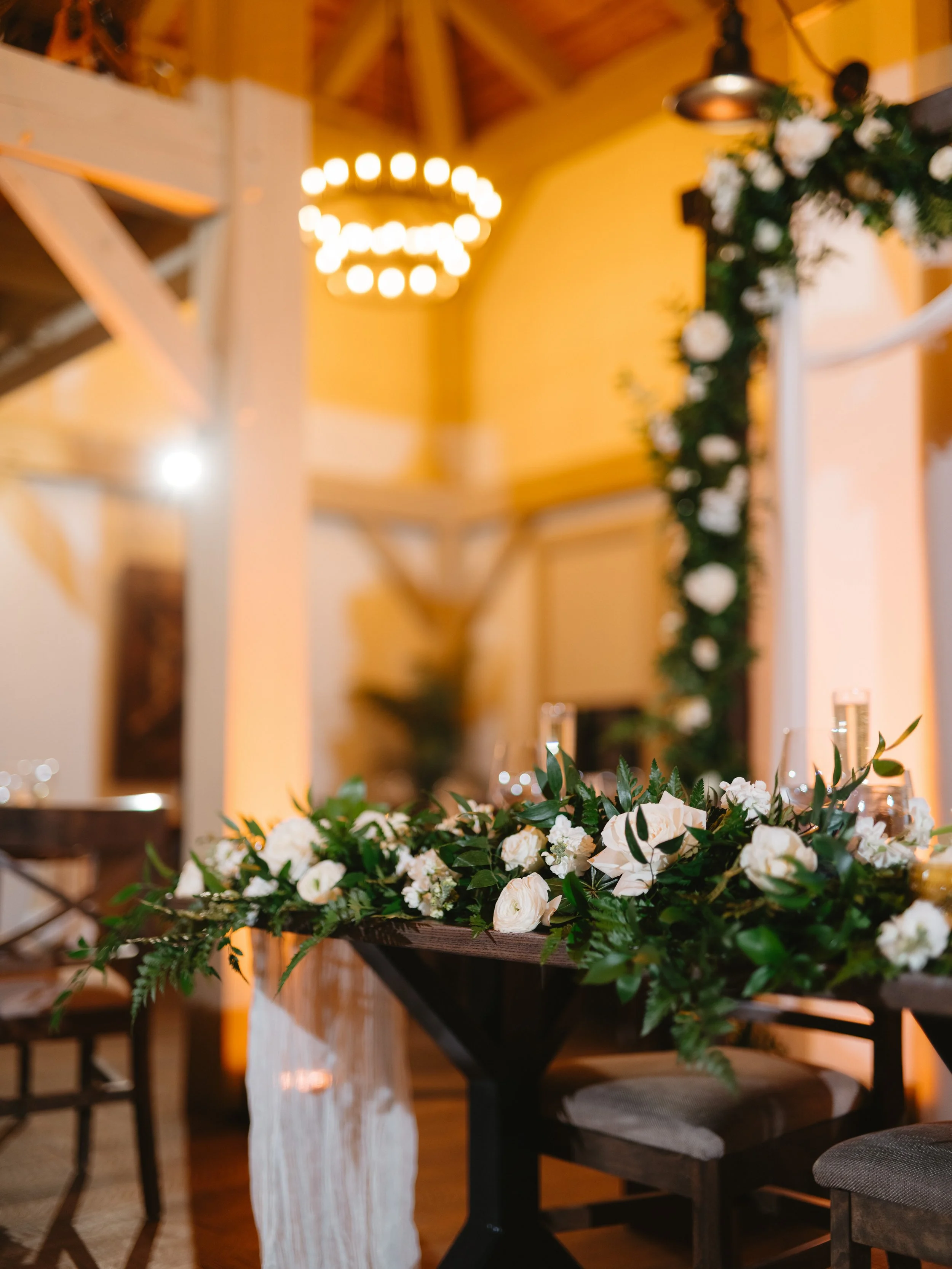A banquet table decorated with a lush green and white floral arrangement, situated in a rustic wooden venue with warm lighting and a modern chandelier.