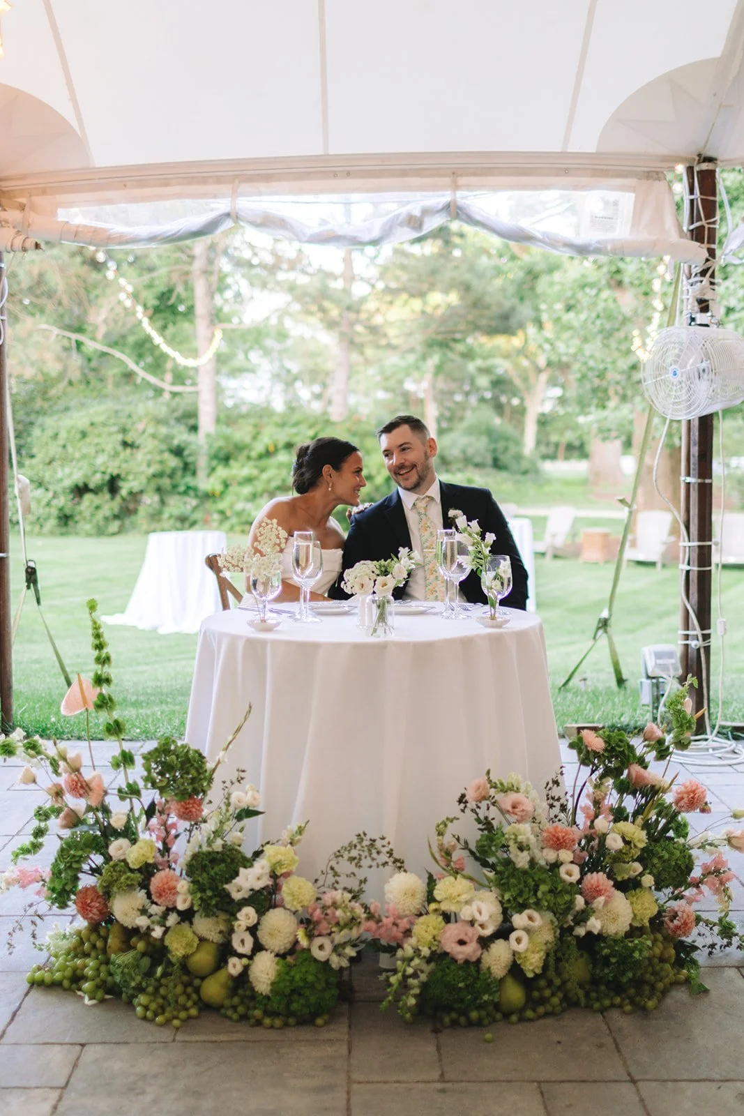 Bride and groom sitting at a table with floral decorations during an outdoor wedding reception.