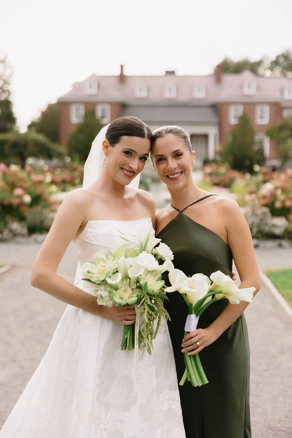 Two women standing outdoors, one in a white wedding dress holding a bouquet of white and green flowers, and the other in a dark green dress holding white calla lilies, smiling at the camera with a large house and garden in the background.