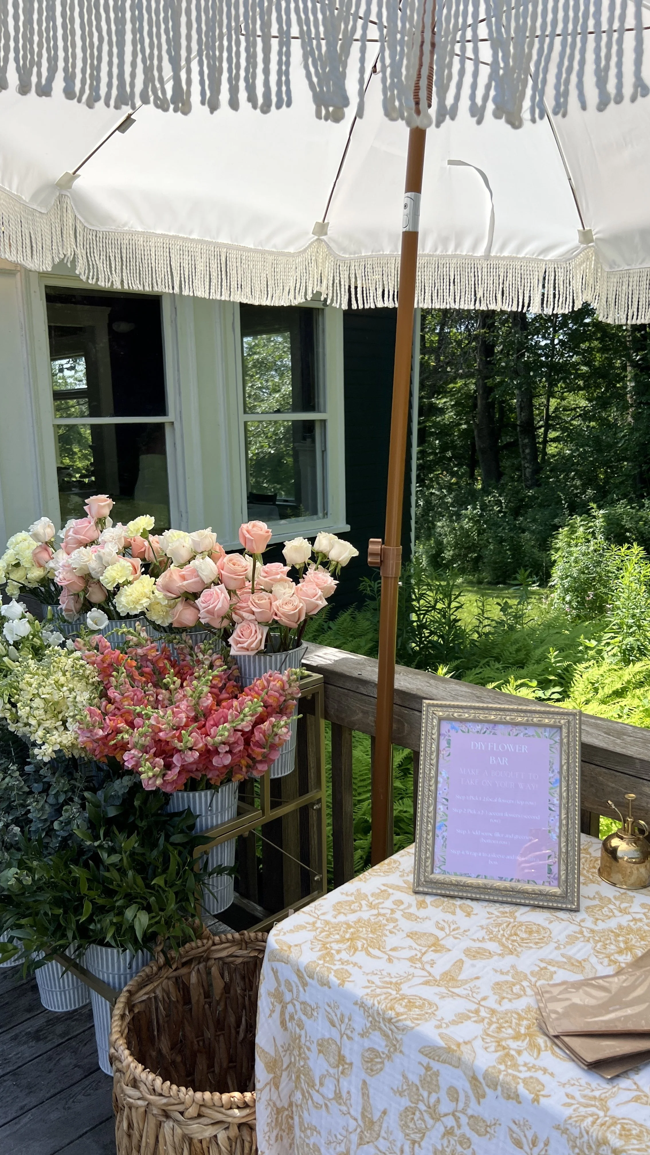 A table with a floral tablecloth under a white patio umbrella, decorated with pink and white roses, and a framed sign for a DIY flower bar.