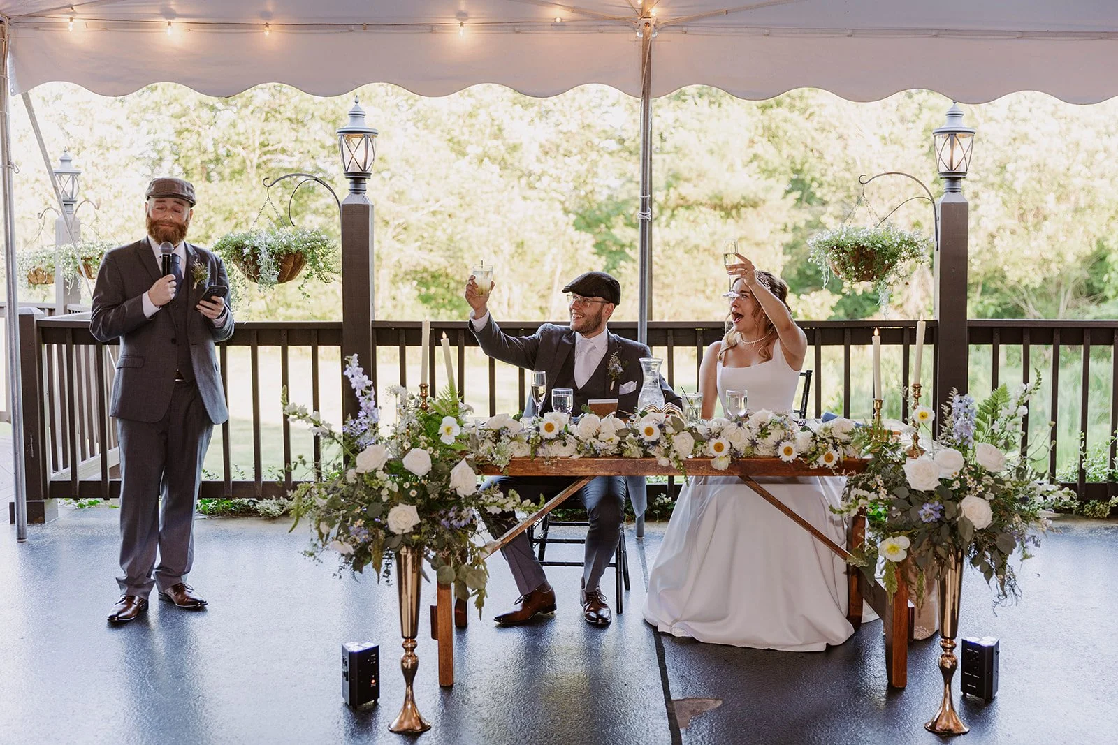 Wedding reception with a bride and groom sitting at a decorated table, raising glasses, and a man giving a speech with a microphone under a tent with string lights and hanging plants, outdoors with trees in the background.